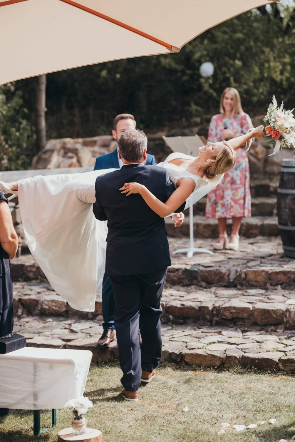 Ein Brautpaar wird bei einer Hochzeit im Freien von einem Mann im Anzug gehalten, die Braut trägt ein weißes Kleid und einen Blumenstrauß in der Hand, im Hintergrund gibt es eine Frau in einem bunten Kleid und eine leere Bar. Es ist sonnig und es gibt einen Baum im Hintergrund.
