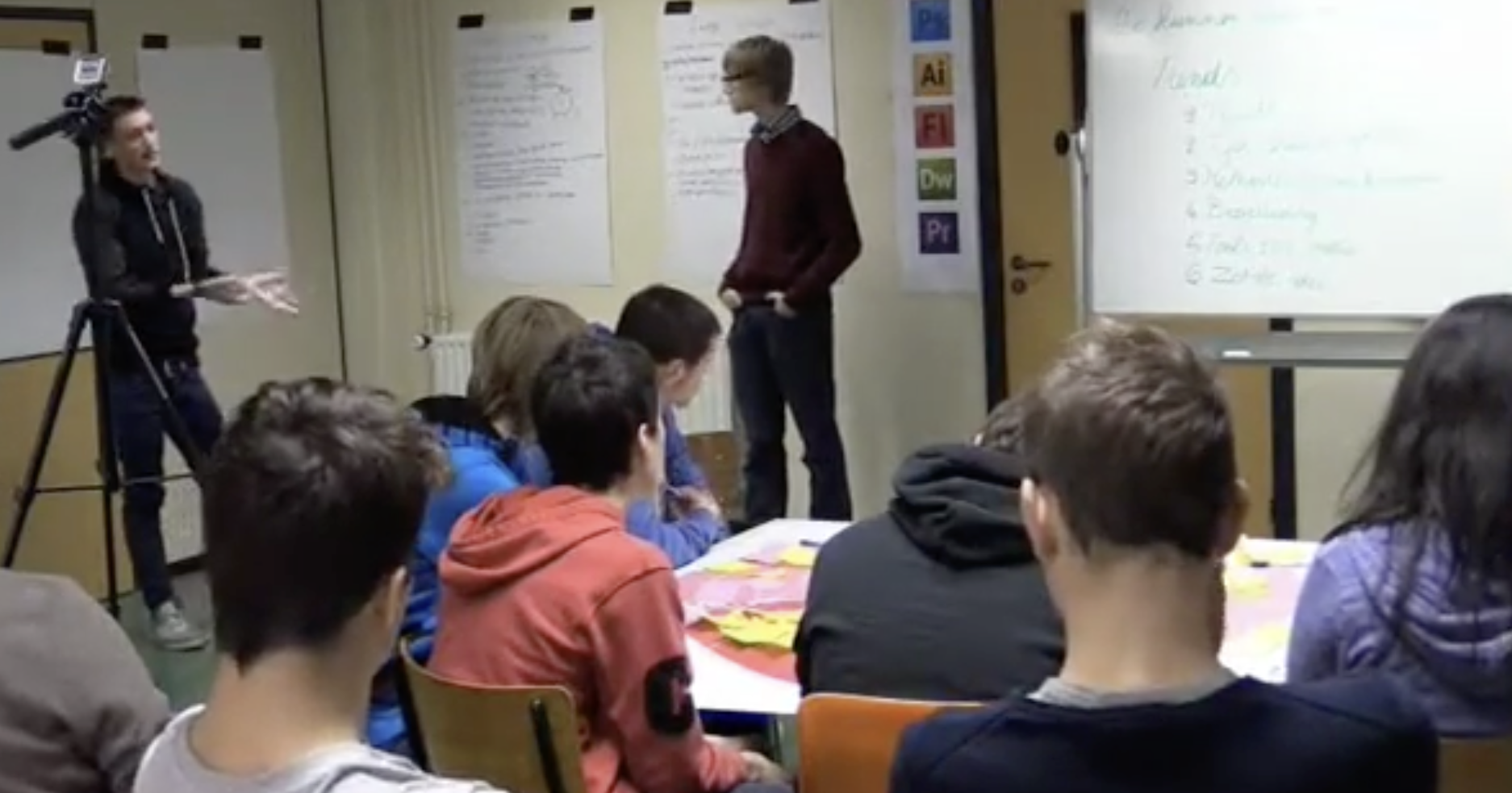 Students attending a classroom presentation with a whiteboard, posters, and a camera operator filming.