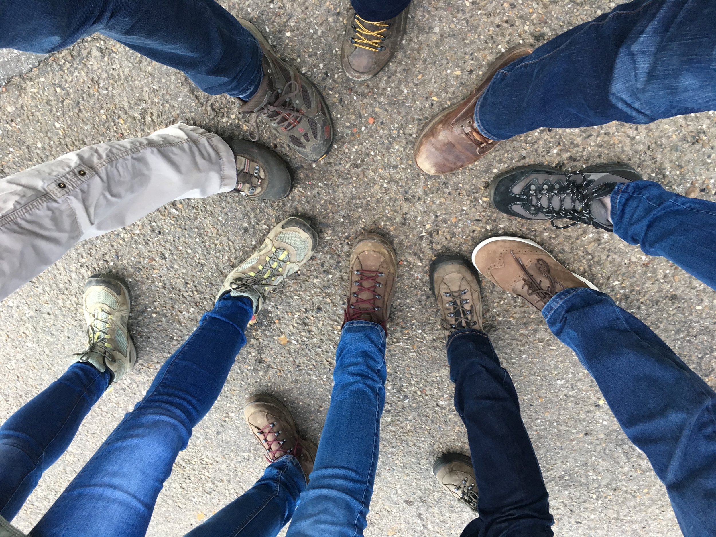 A ground view of eight people standing in a circle, each wearing different styles of hiking shoes and blue jeans or khakis on a gravel surface.