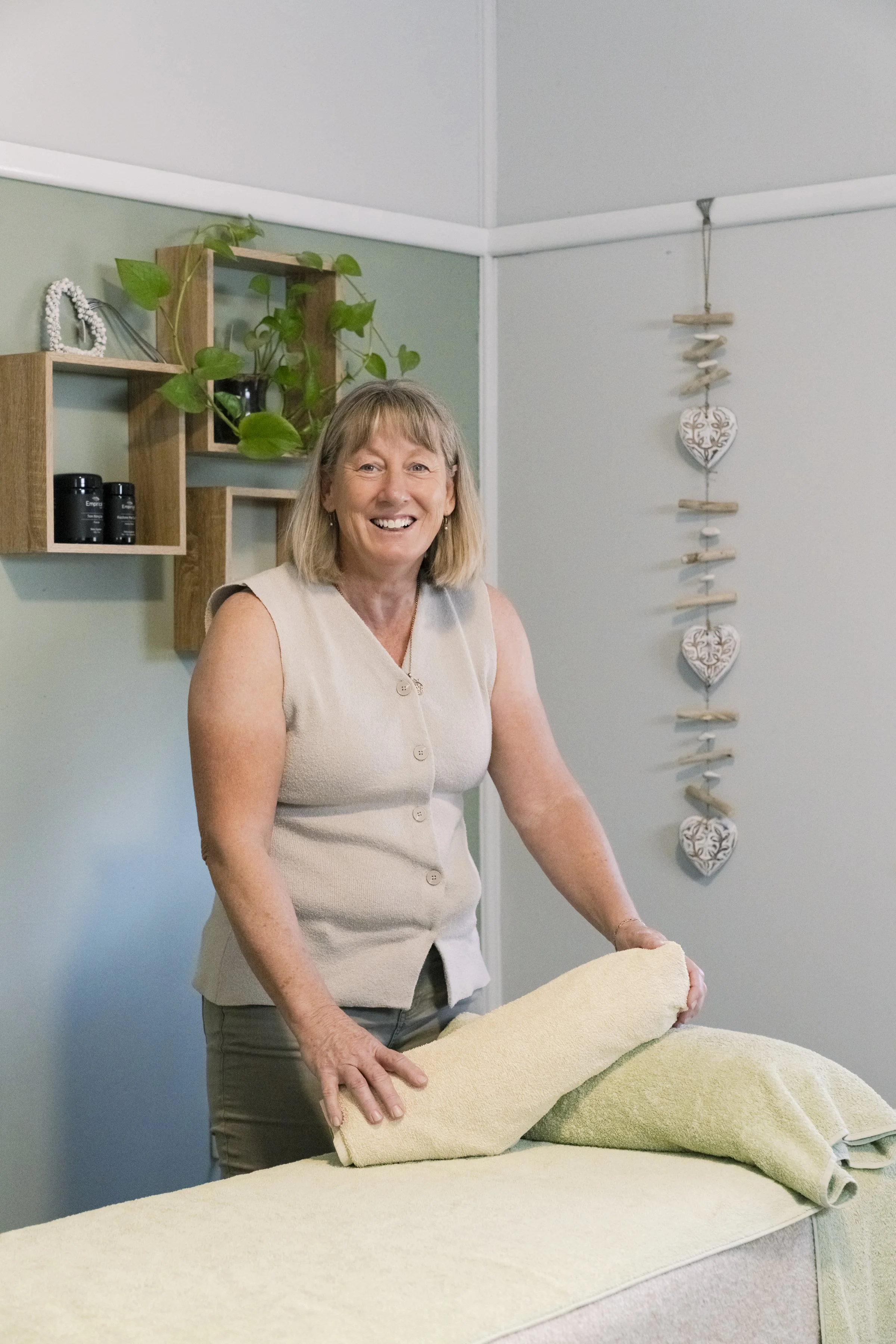 A woman smiling while preparing a massage table with a rolled towel in a spa or massage room.