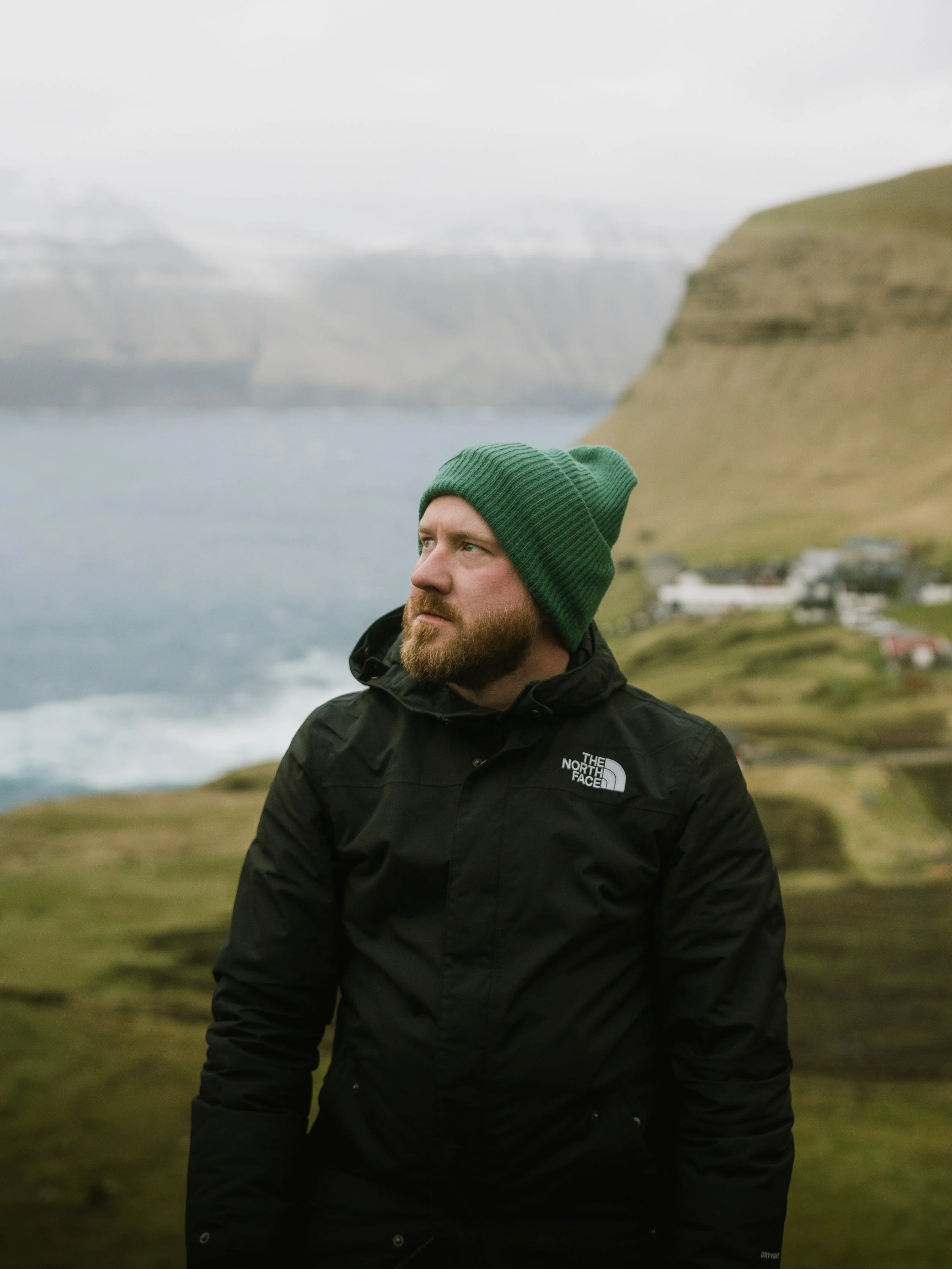 A man with a beard wearing a green beanie and a black North Face jacket standing outdoors with rolling hills, water, and distant mountains in the background.