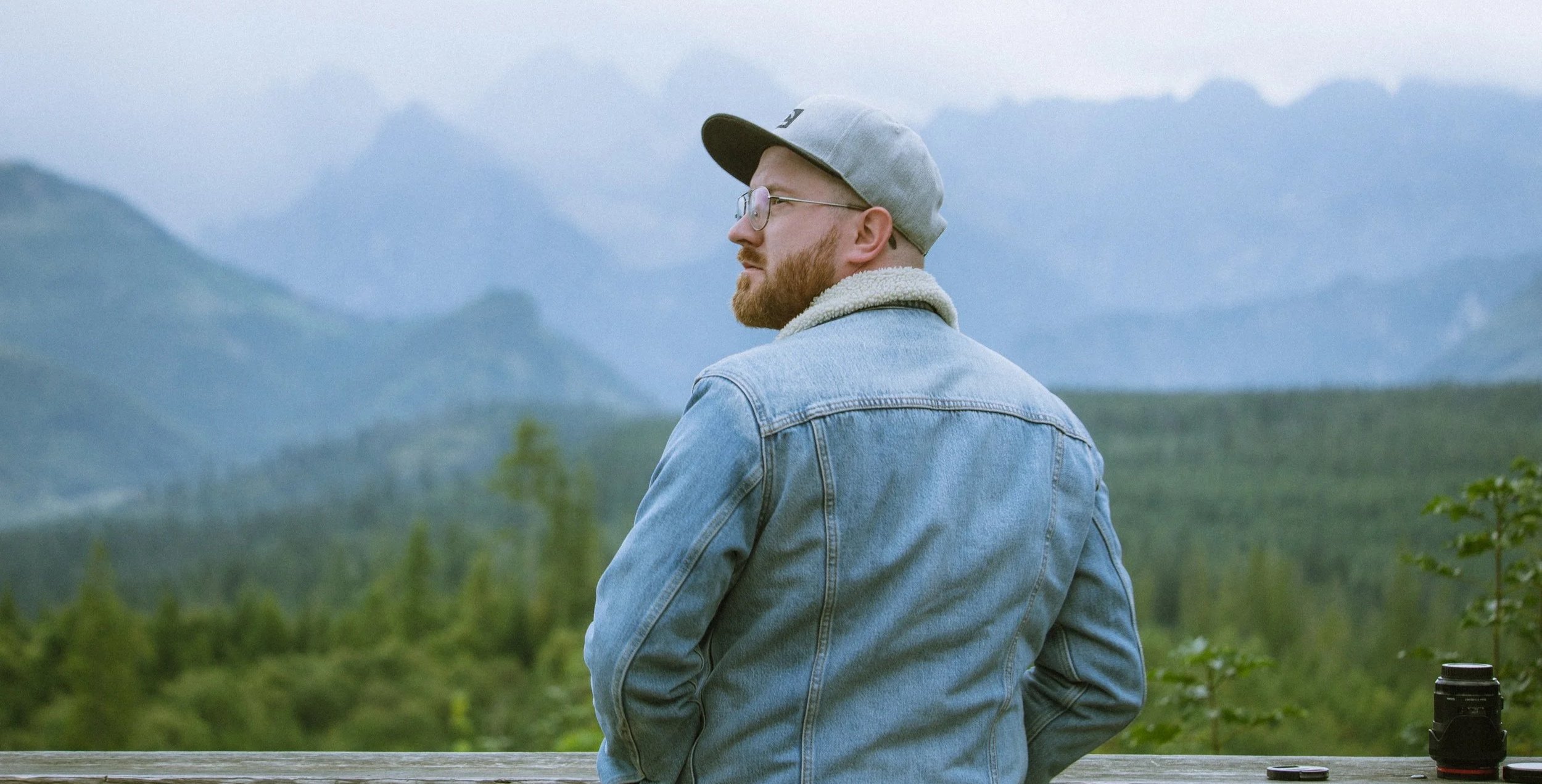 A man with a beard and glasses, wearing a grey cap and denim jacket, stands outdoors overlooking a landscape of mountains and greenery.