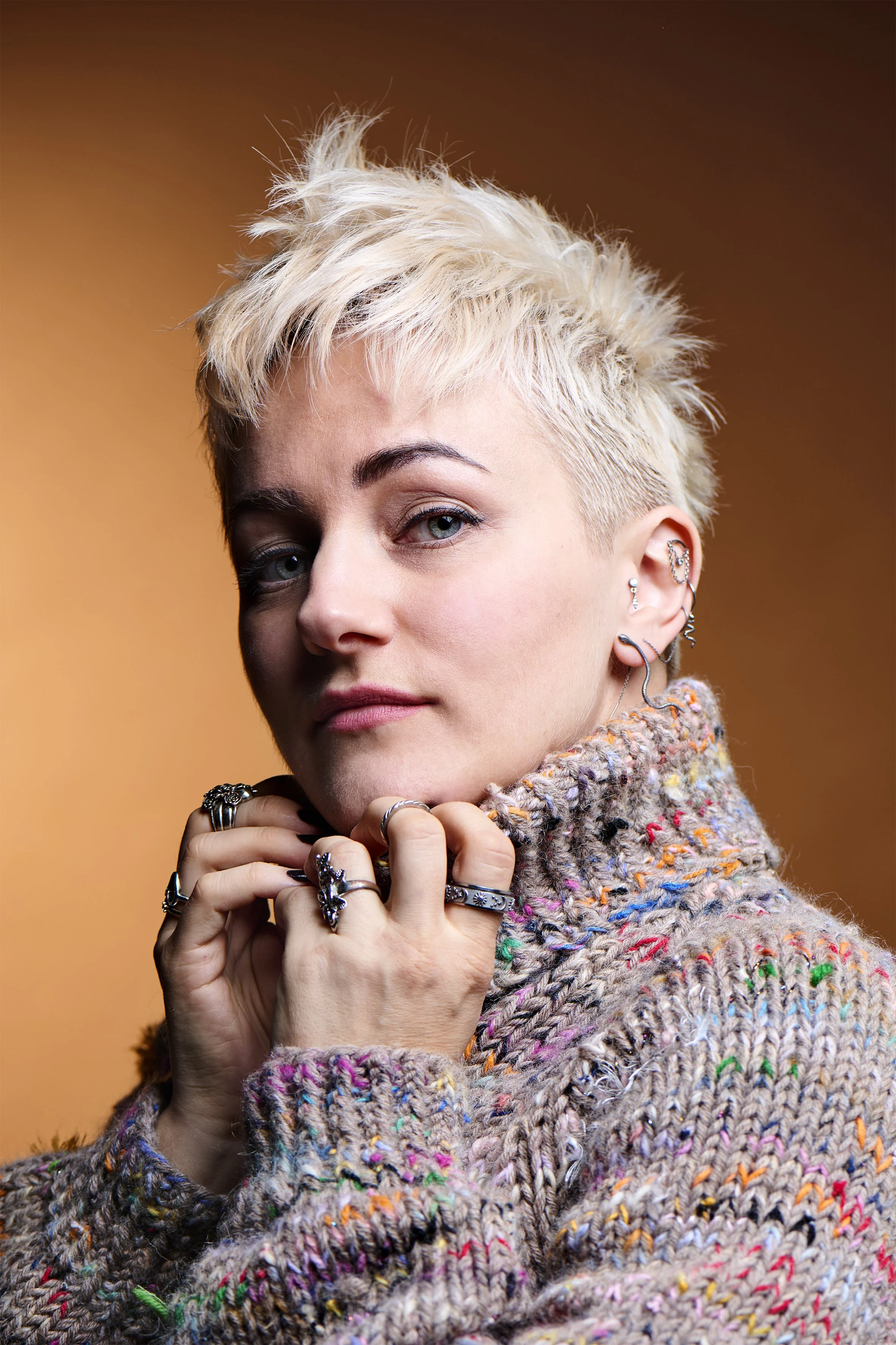 Portræt af en kvinde med kort, blond, skandinavisk look, iført en farverig, strikket sweater, smykker i øret og hænderne tæt på ansigtet, mod en varm, brun-orange baggrund.