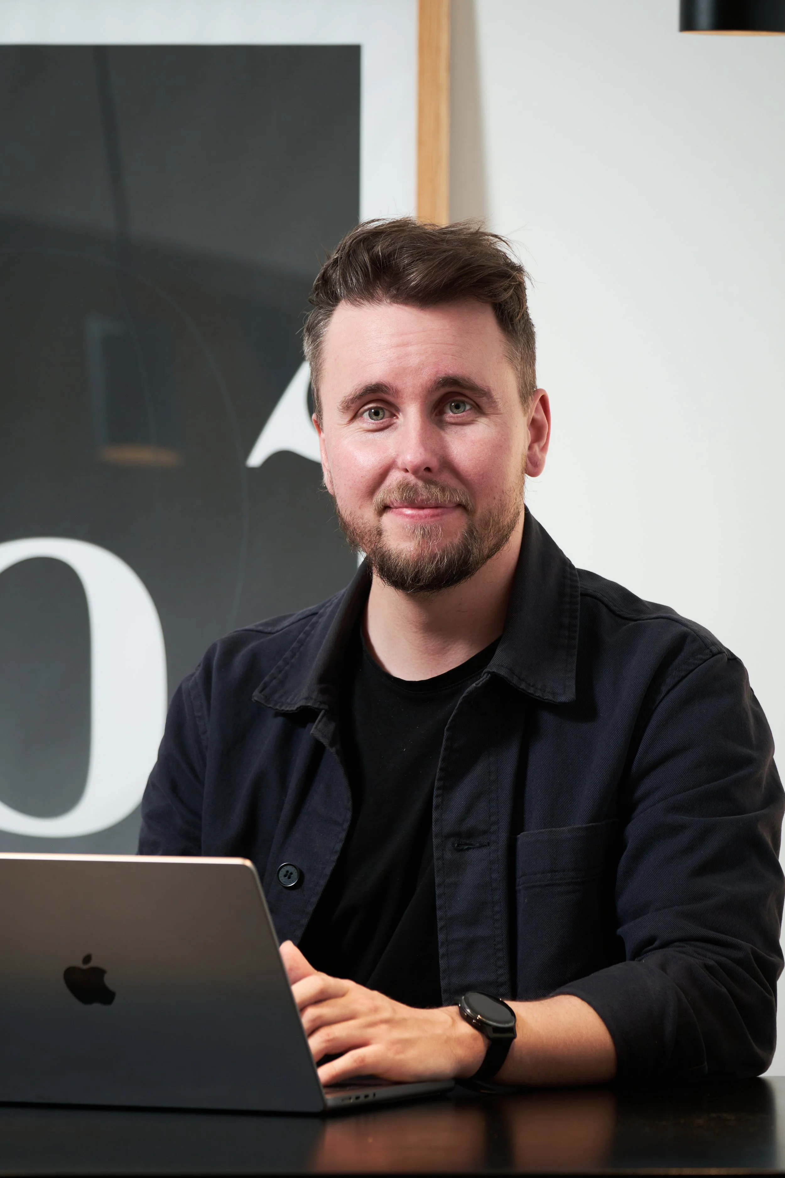 Mand sidder ved en laptop, iført sort skjorte og smartur, med et smilende ansigt, i et moderne kontor.