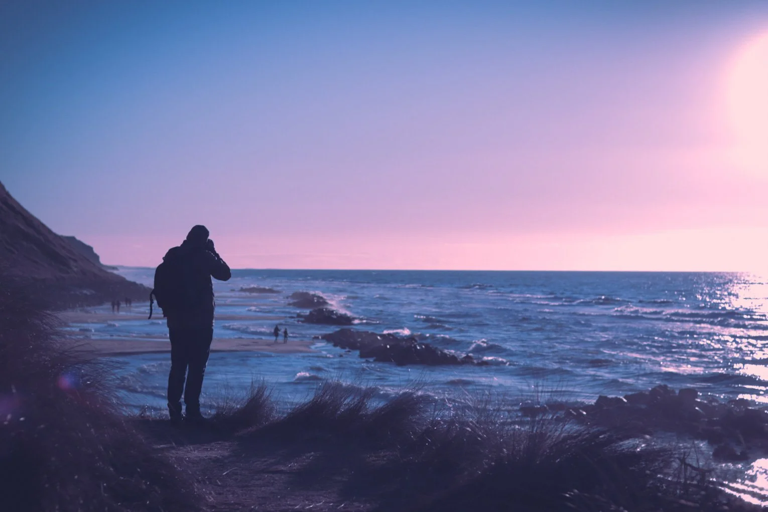 En person står på en strand med klitter, og ser ud over havet ved solnedgang eller solopgang, med en lilla og lyserød himmel.