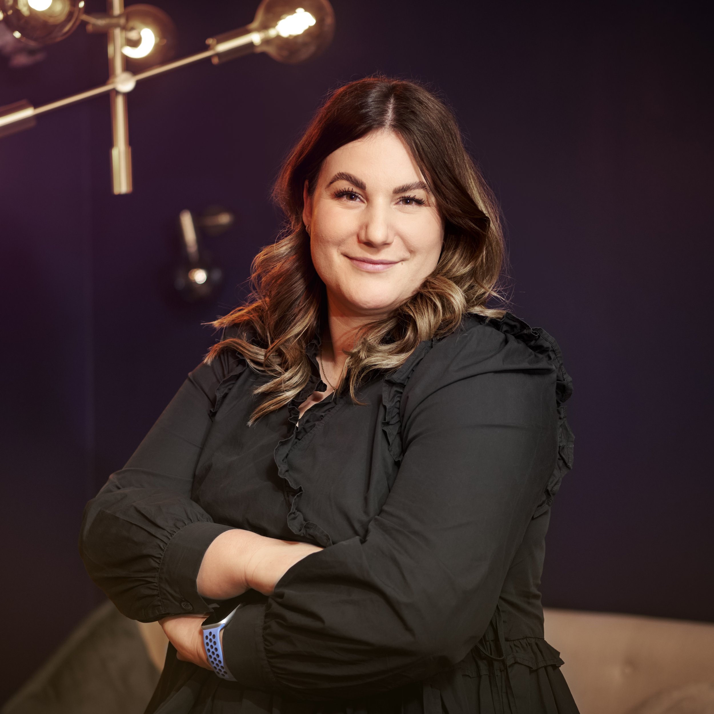 En ung kvinde med langt, bølget brunt hår, smilende, iført sort jakke, står med armene krydsede, bag den mørke baggrund.
