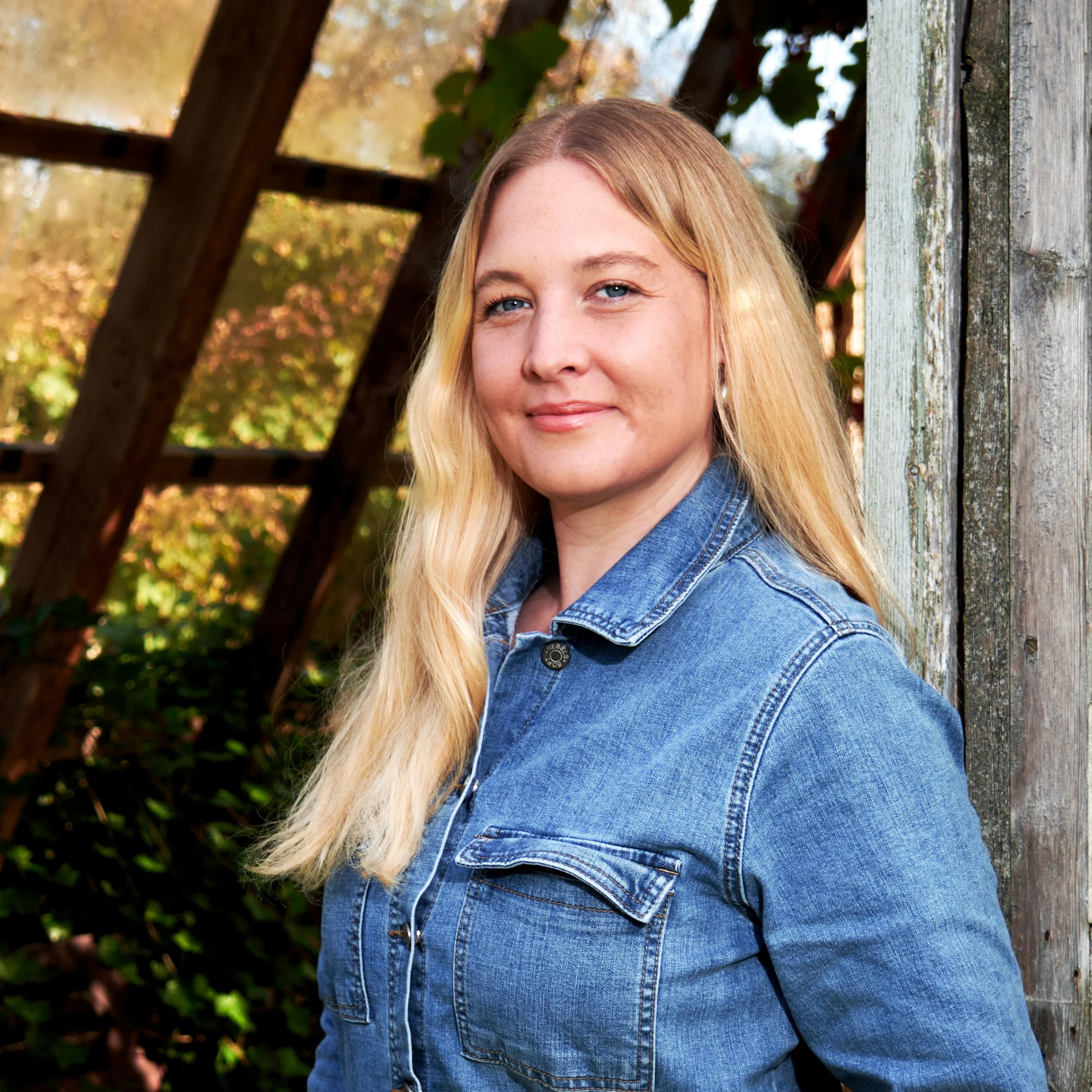 Nærbillede af en ung kvinde med blondt hår, blå øjne og smil, der står ved en gammel trægyld til udendørs, med efterårsløv i baggrunden.