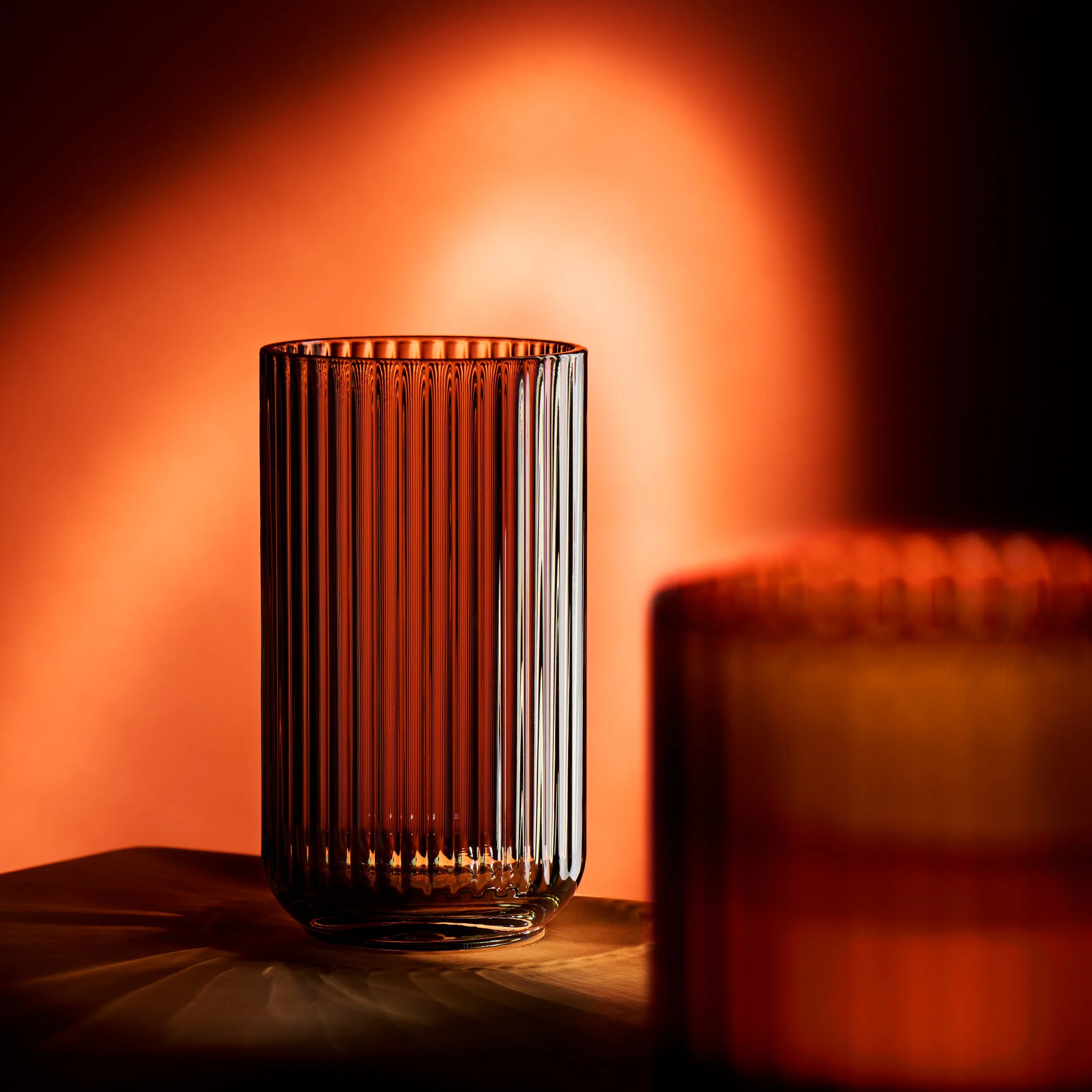 Et tomt glas med vertikale ridser står på en træoverflade, med en varm, orange baggrundslys, der skaber skygger på overfladen.