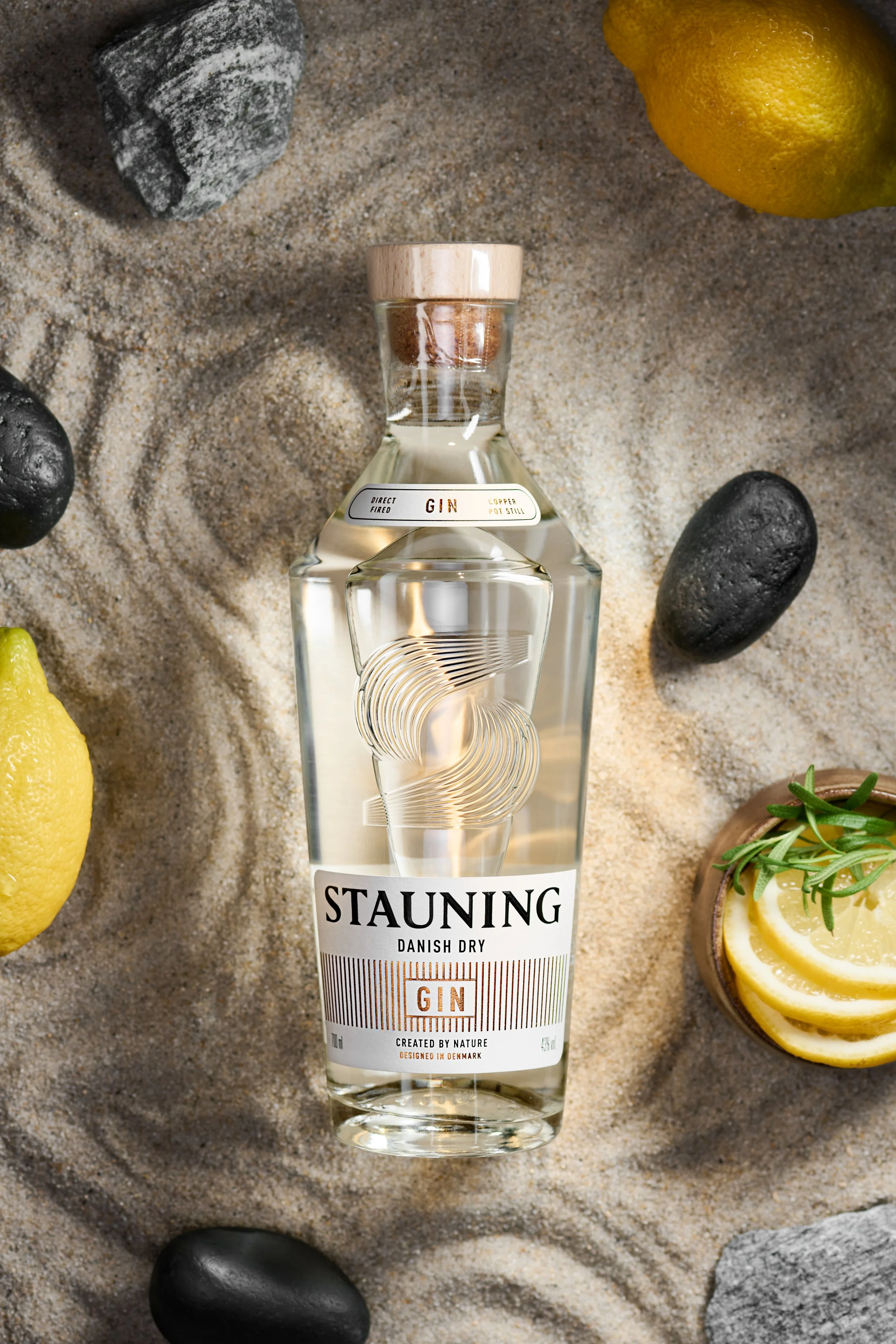 Et gennemsigtigt glas med gin, omgivet af citronbåde, sorte og grå sten, og en skål med citron- og rosmarinekviste på sandet.