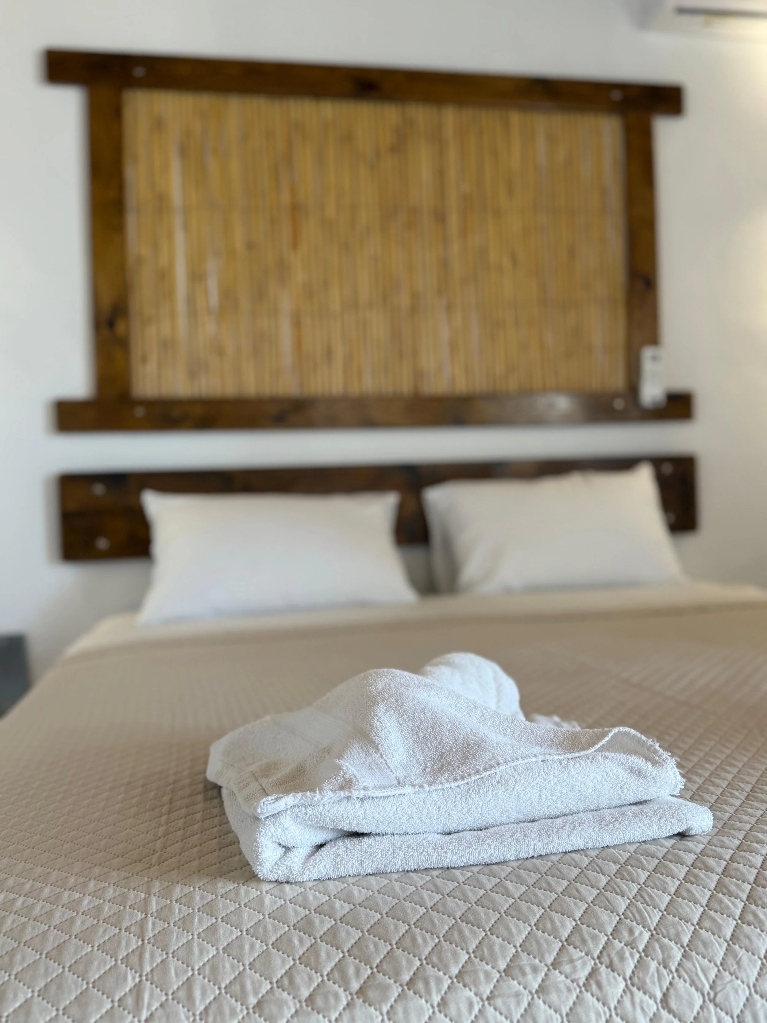 A bed with a folded white towel and a smaller towel on top, positioned in front of a wooden headboard with a bamboo pattern.