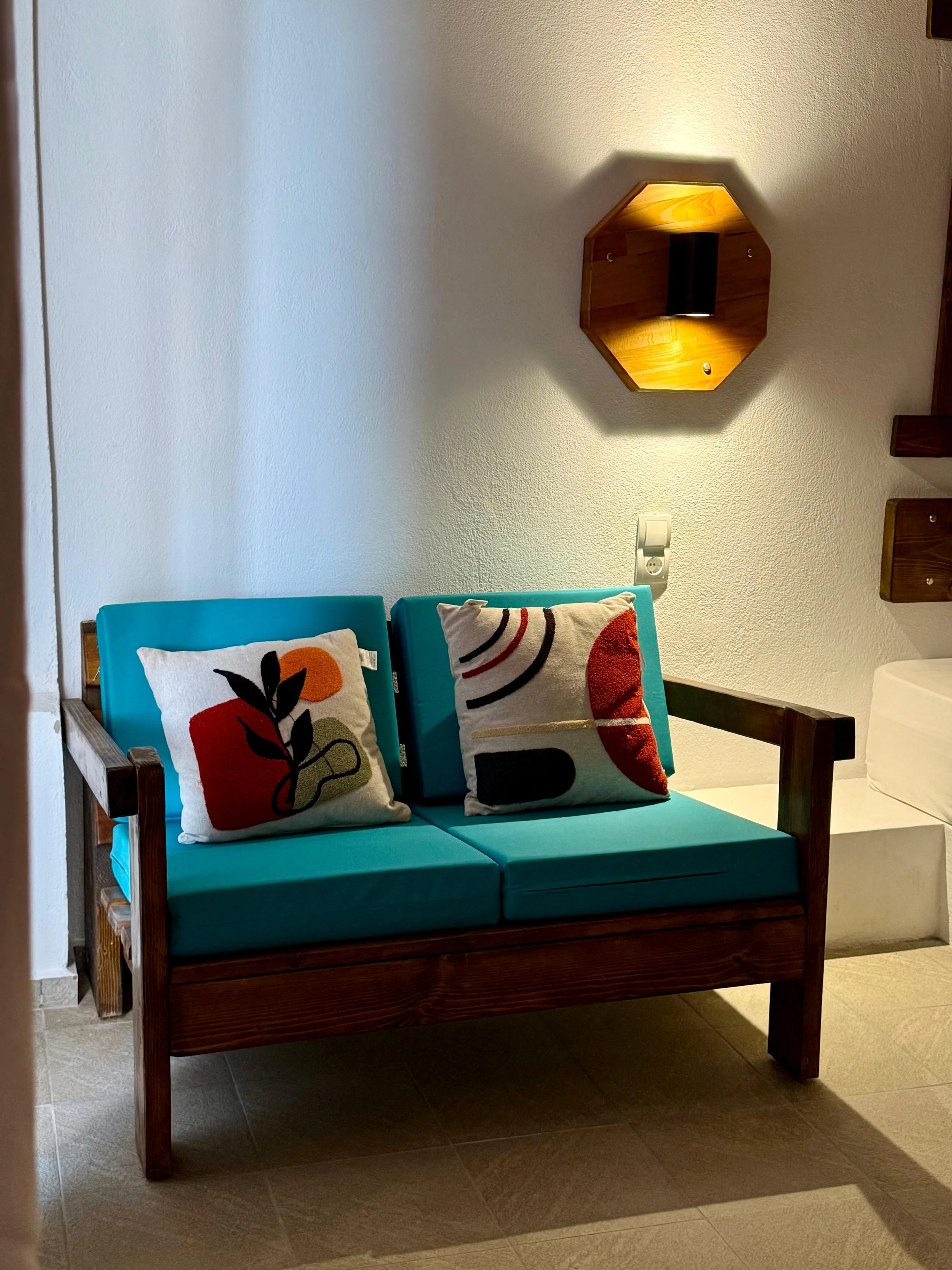 A wooden bench with teal cushions and two decorative pillows with abstract designs, against a white wall with a wall-mounted lamp and electrical outlet.