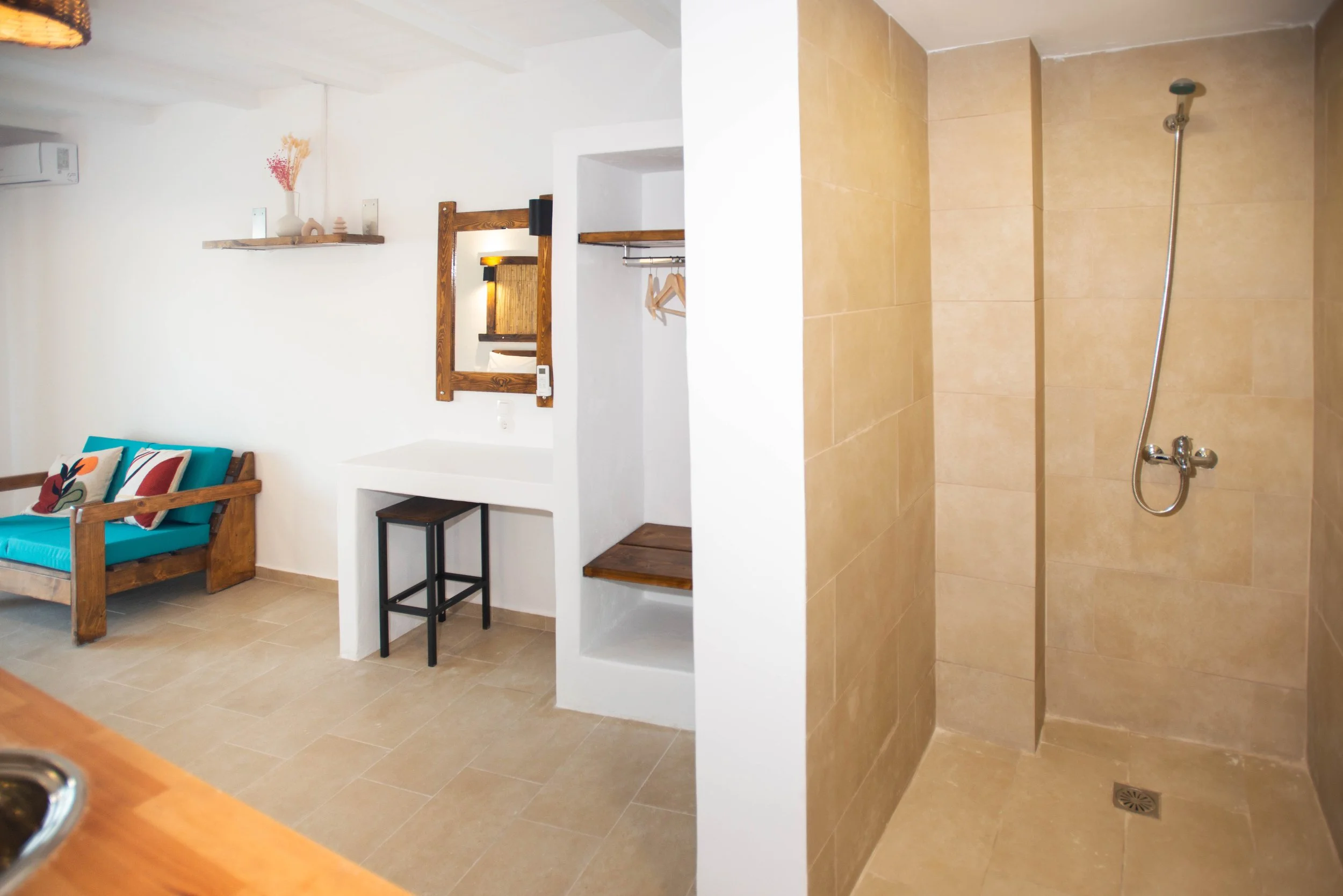 Interior of a small room with a bed, a mirror, shelves, and a shower area with beige tiles, a handheld showerhead, and a floor drain.