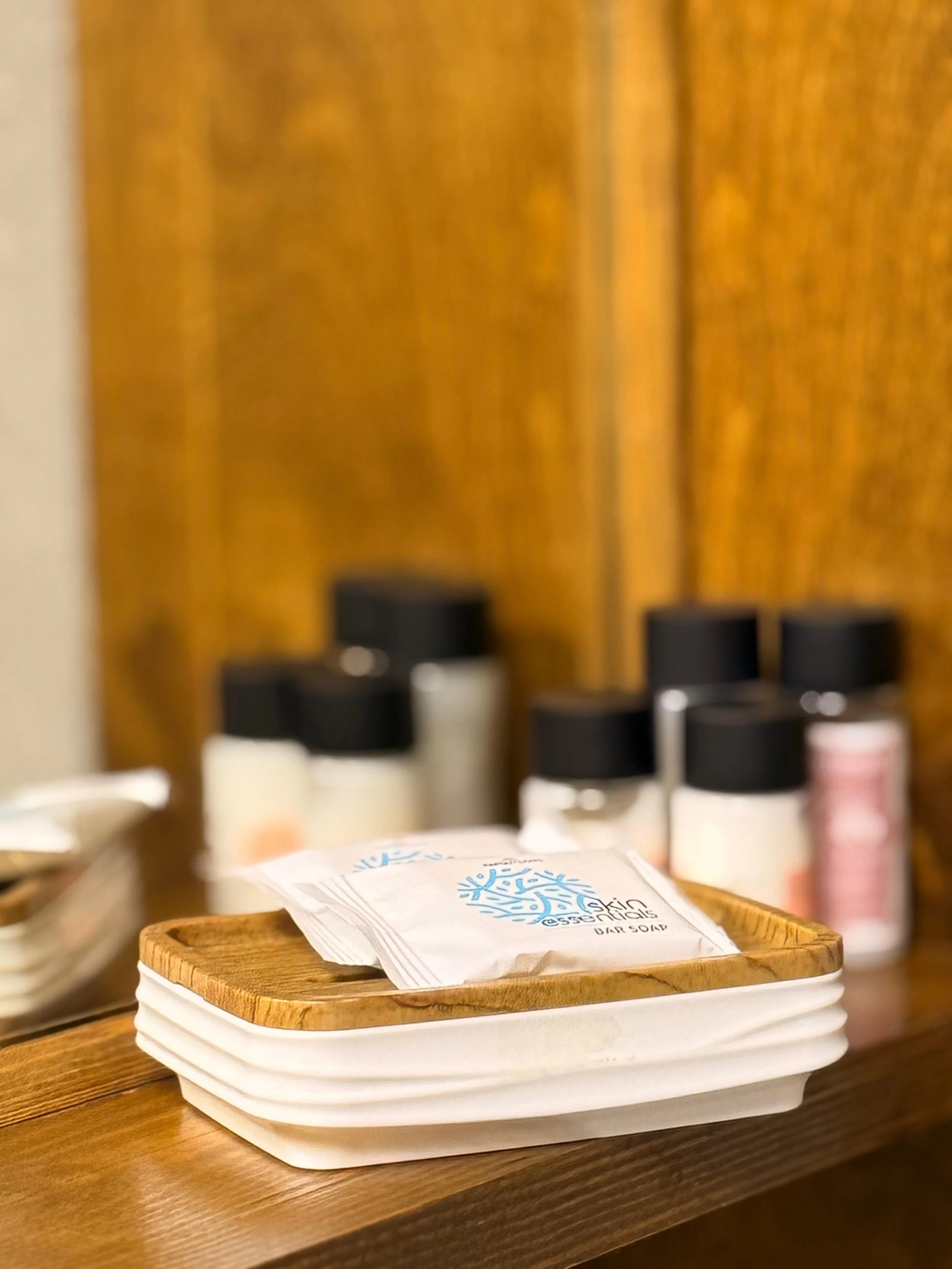 Close-up of a wooden tray with white paper napkins featuring a blue logo or text, placed on a wooden countertop. In the background, out-of-focus bottles and containers are visible.