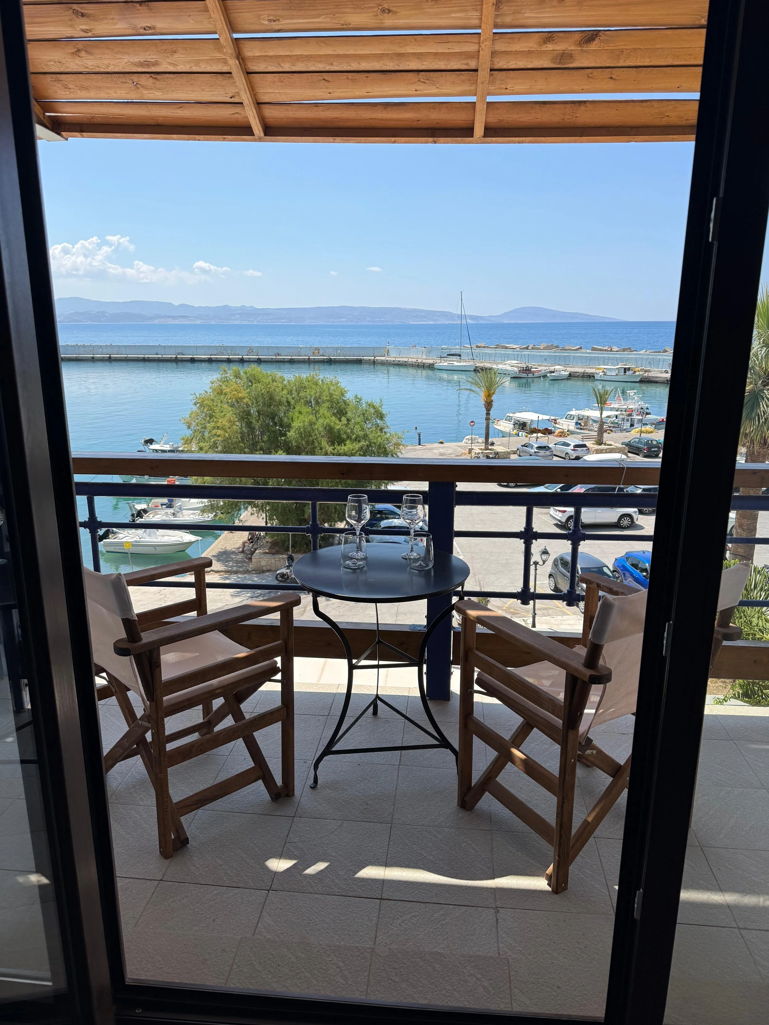 A balcony with a small round table set with two wine glasses, surrounded by three wooden chairs, overlooking a harbor with boats and a view of the ocean and distant mountains.