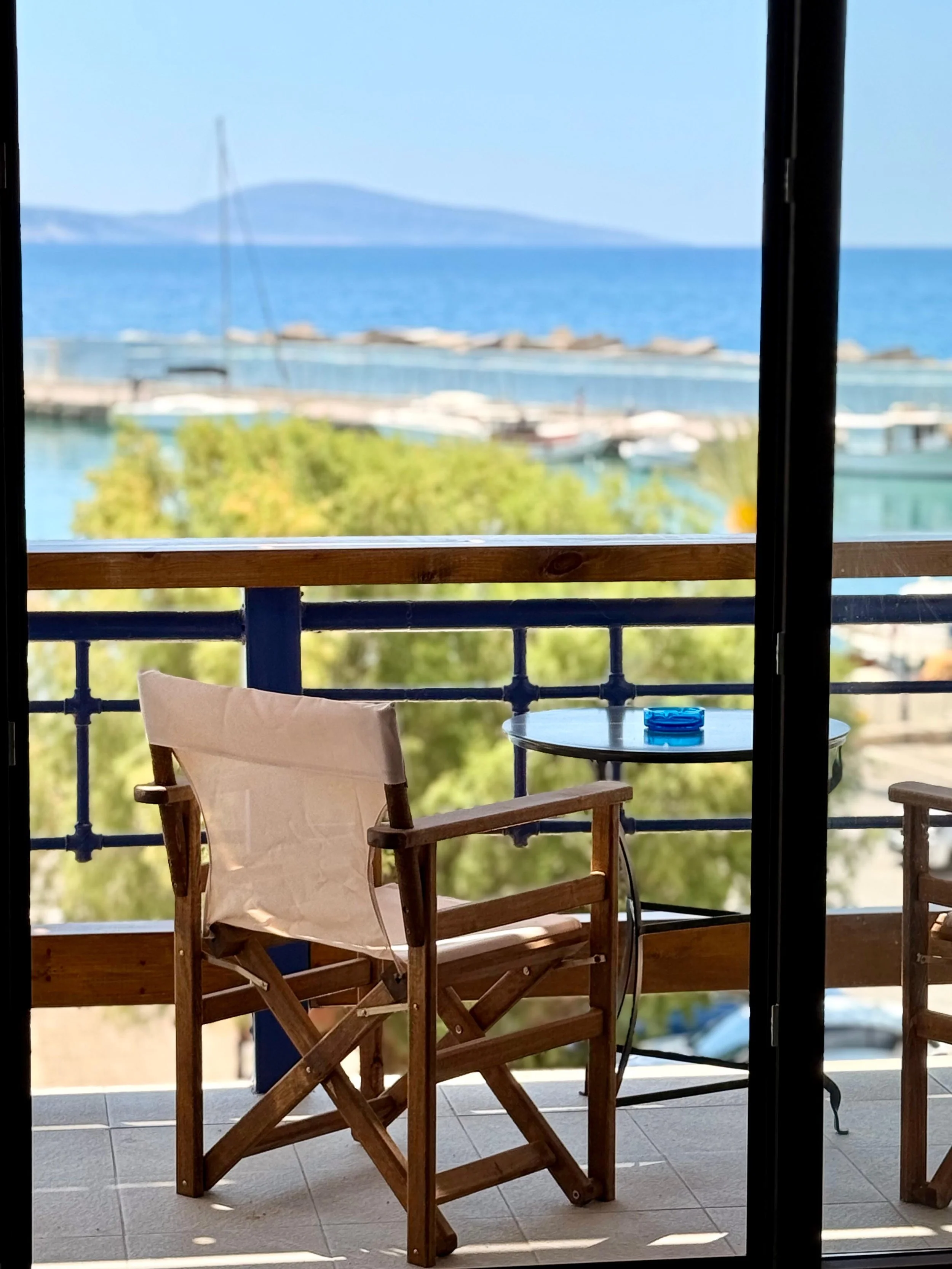Balcony with wooden chairs and a small table, overlooking Agia Galini marina with boats and a distant island, under a clear blue sky.