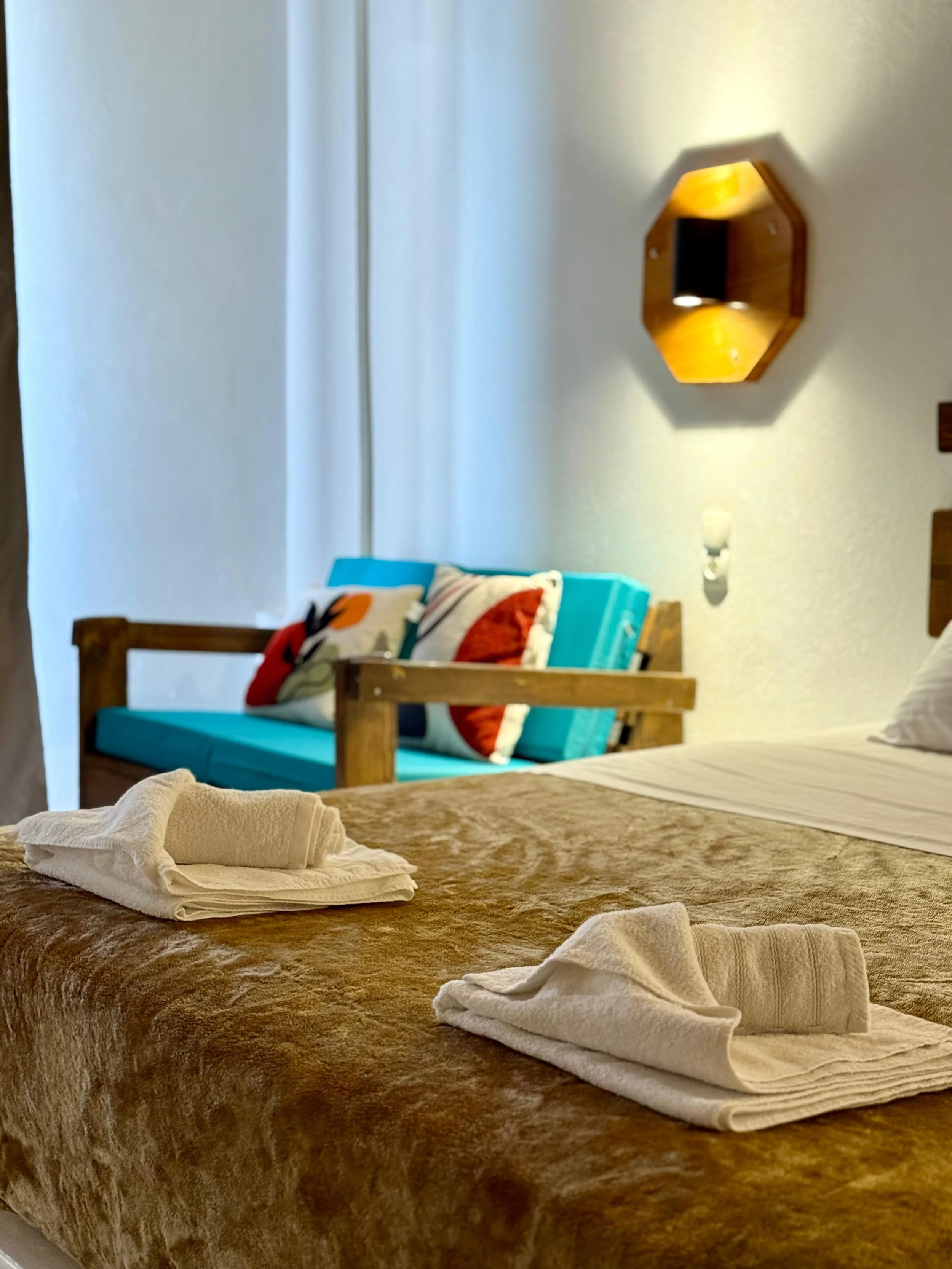 A hotel room with a bed covered in a brown textured bedspread, with neatly folded white towels on top. In the background, there's a wooden bed frame, a turquoise mattress, and pillows with decorative cases. The wall has a wooden shelf with a mirror a