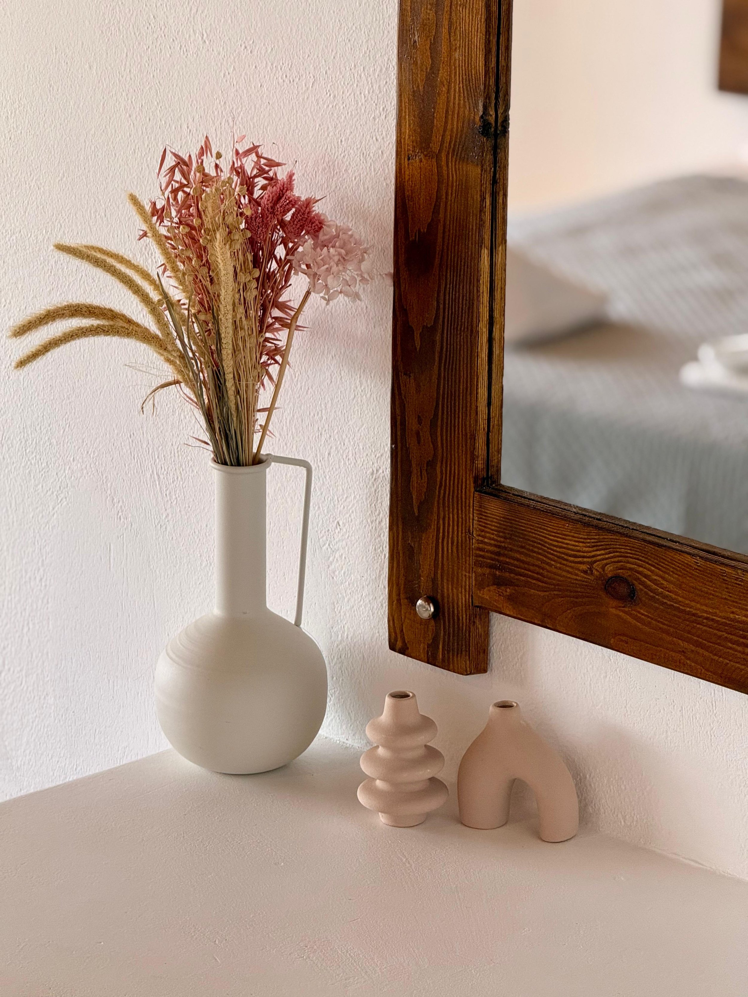A white vase with dried pink and beige flowers is placed next to three minimalist pink ceramic vases of varying sizes and shapes, with a wooden-framed mirror hanging on the white wall behind them.