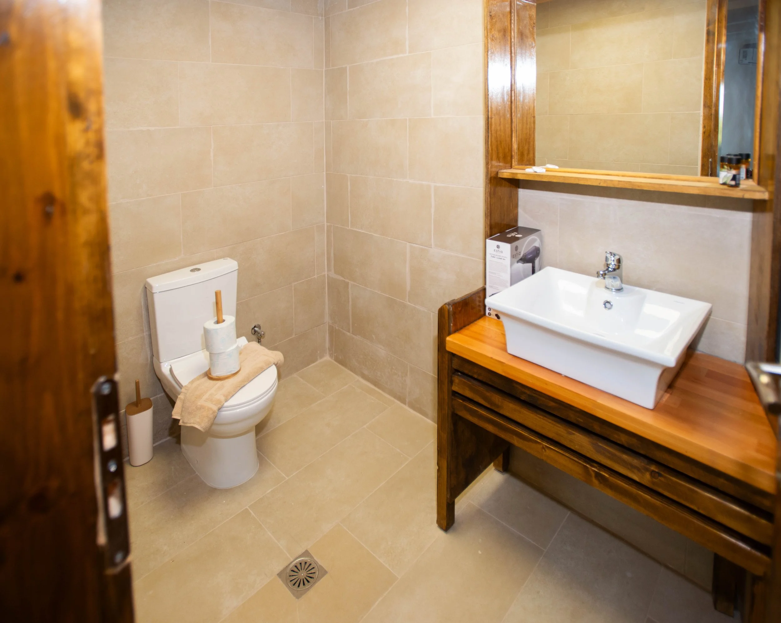 A small bathroom with beige tiled walls and floor, a white toilet with a wood toilet brush next to it, a bar of soap on the top of the toilet tank, a large rectangular mirror above a wooden shelf, and a white sink with a silver faucet. There are smal
