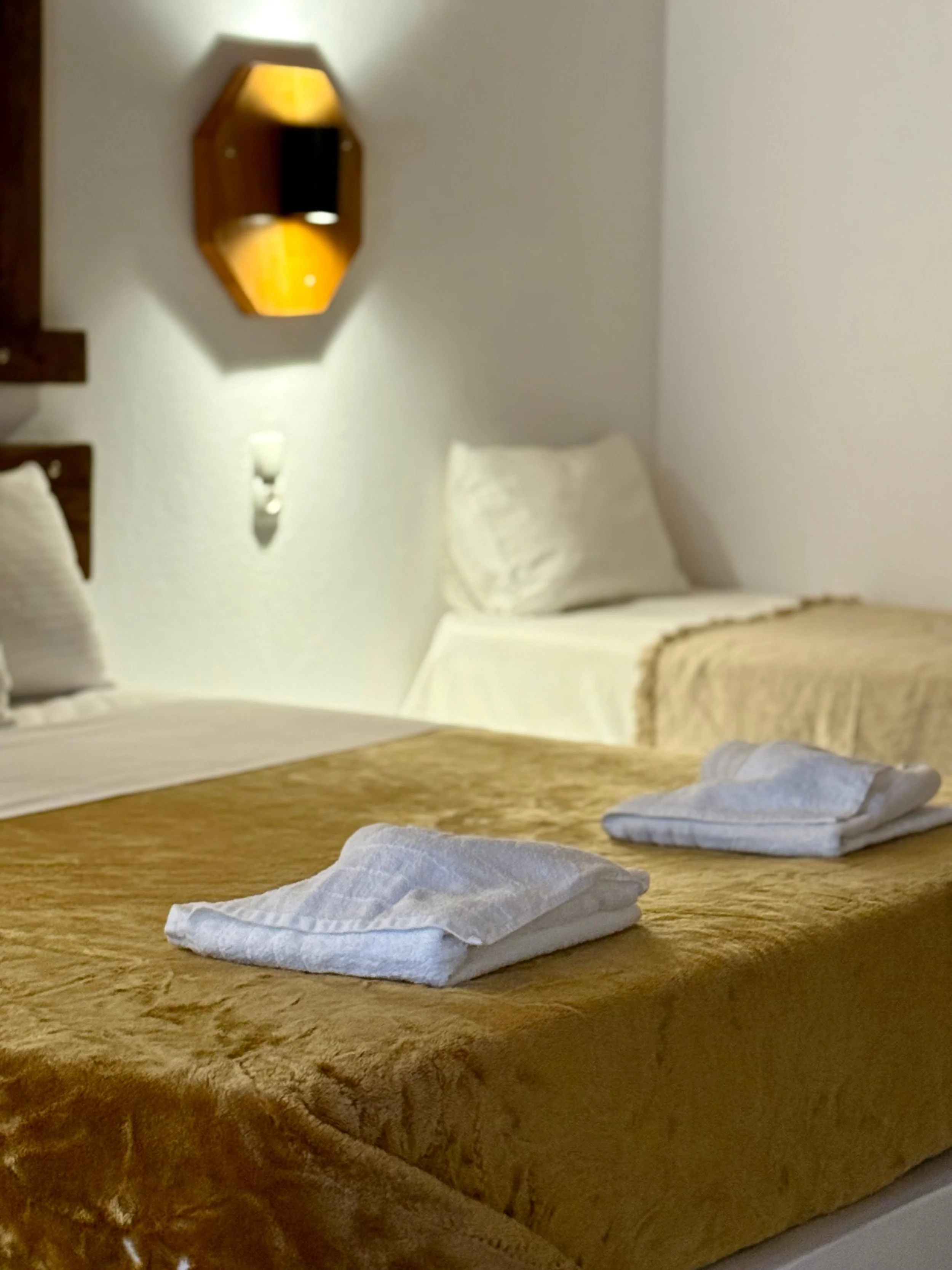 A cozy bedroom with a bed that has white linens and a wooden headboard. There are two white pillows on the bed. Two folded gray towels are placed on a brown, fluffy blanket at the foot of the bed. A wall-mounted lamp provides soft lighting, and a dec