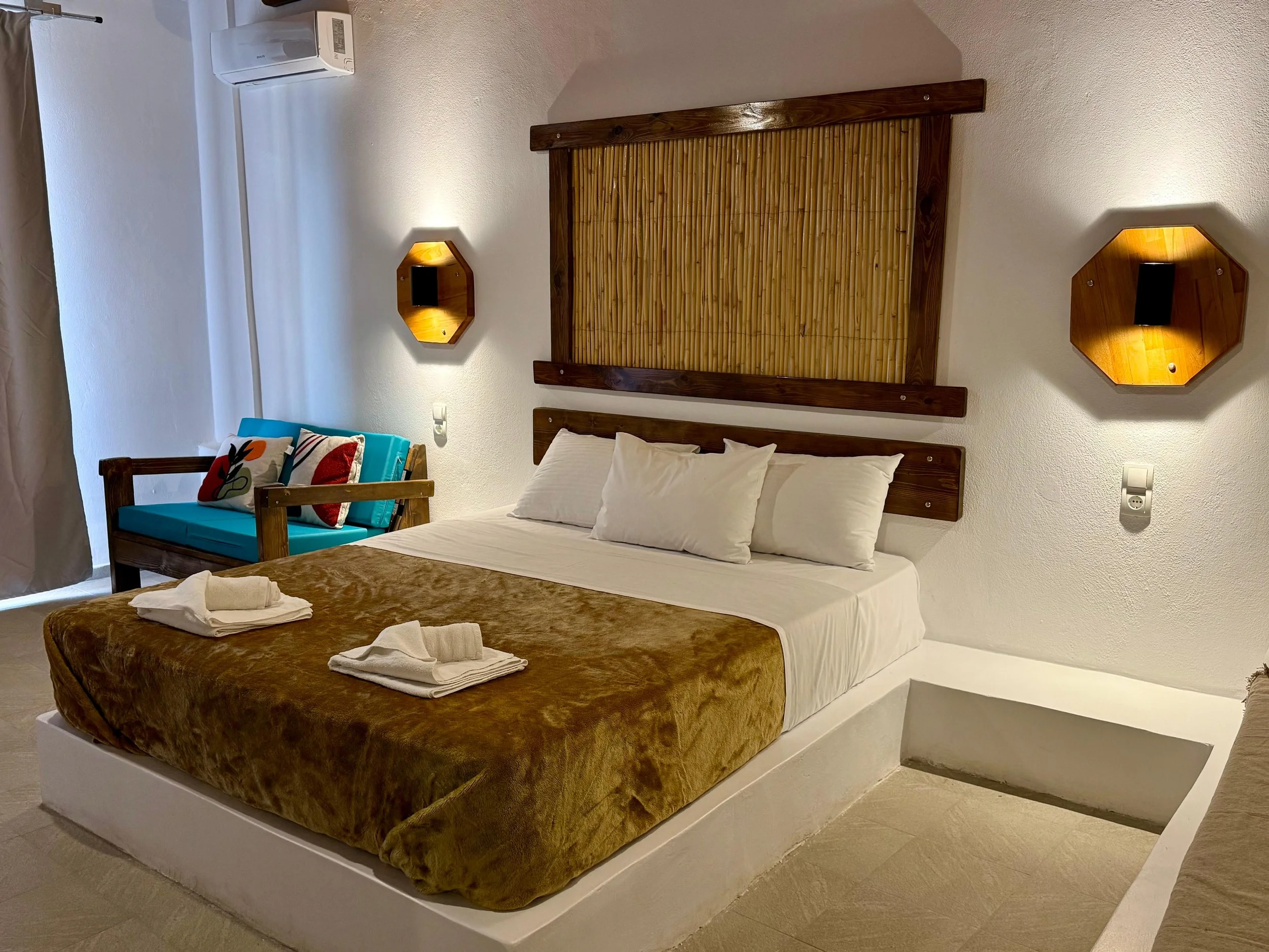 A hotel room with a large bed covered in a brown velvet bedspread, two white pillows, and folded towels. There is a wooden headboard and two wall-mounted lamps with yellow shades. To the left, there is a sitting area with a turquoise cushioned bench 