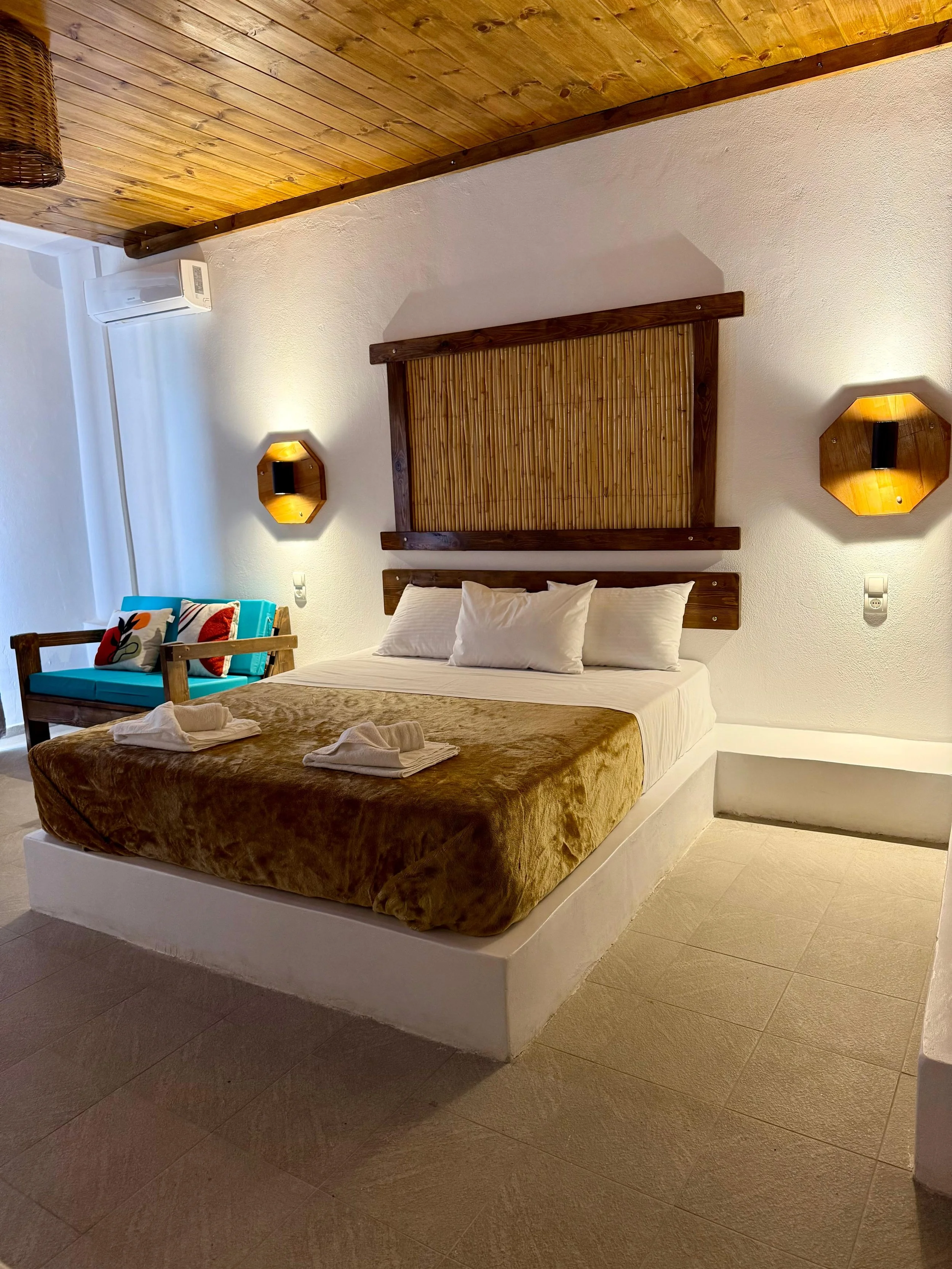 A bedroom with a wooden ceiling and white textured walls. There is a large bed with a brown velvet blanket and white pillows. Two wooden wall-mounted lamps with yellow light are on either side of the bed. A wooden headboard with bamboo decor is behin