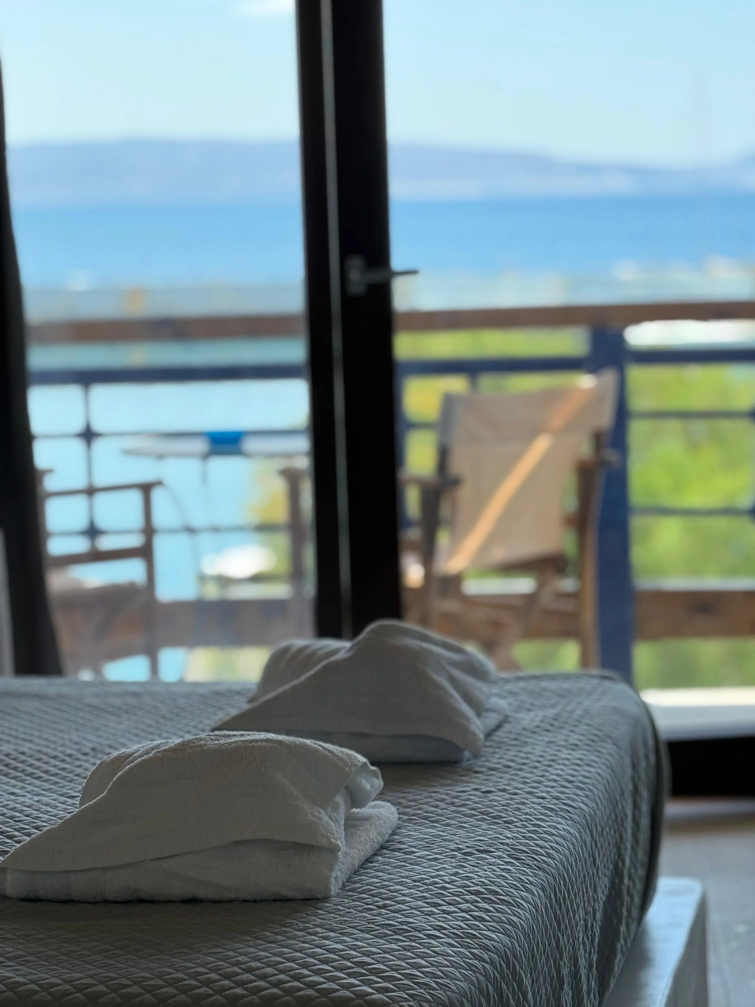 Interior view of a bedroom with a bed, blanket, and pillow in the foreground. A sliding glass door opens to a balcony with a table and chairs, overlooking a blue body of water and distant land.