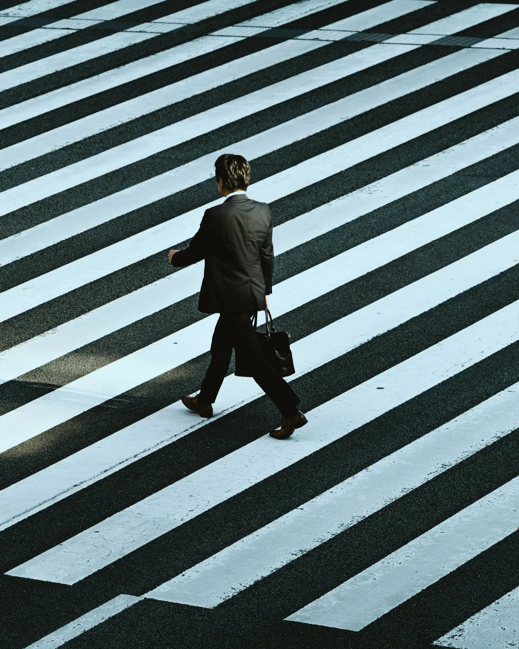 A man in a black suit carrying a black handbag crossing a black and white striped crosswalk.