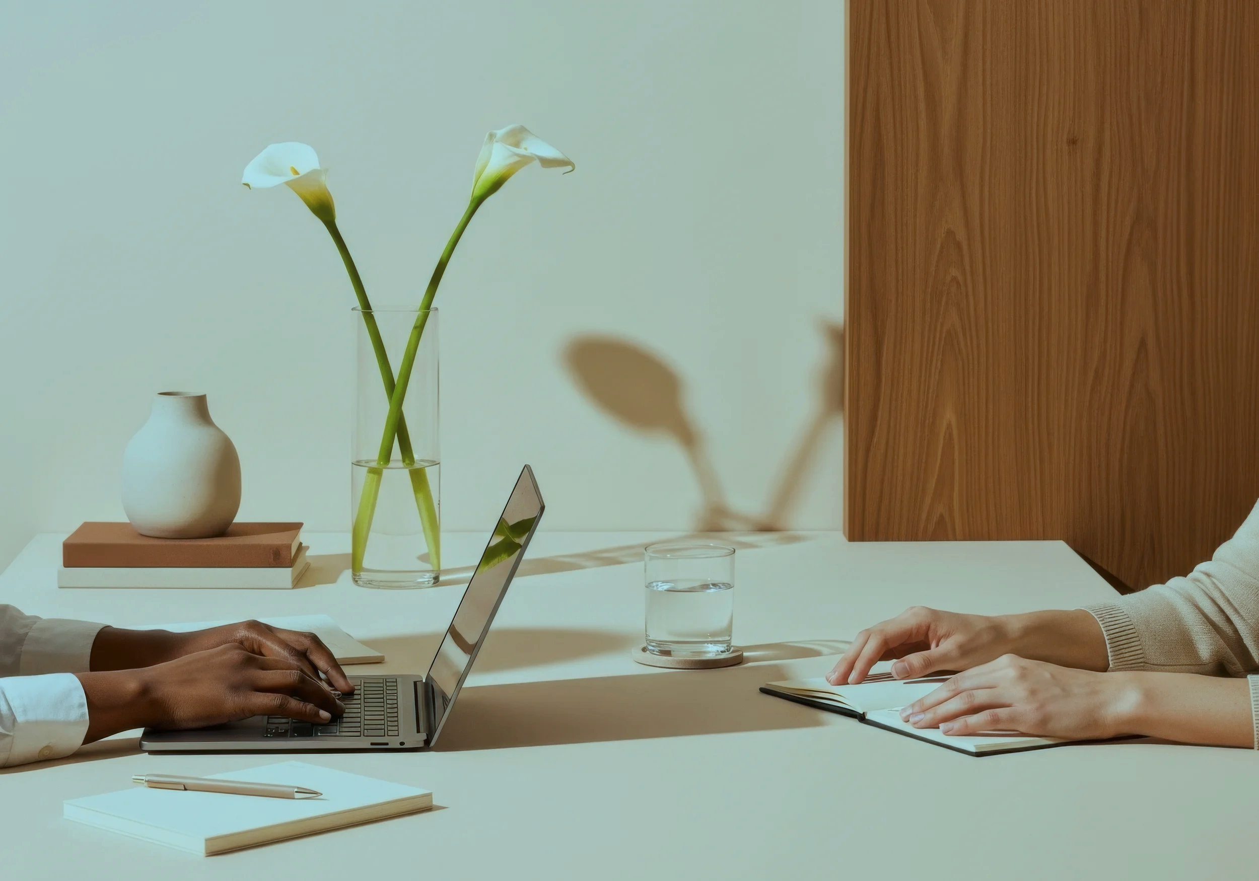 Two people sitting at a light-colored table, working on laptops. A white vase with two calla lilies, a closed book, a notebook, a pen, and two glasses of water are on the table. A wooden panel is on the right side of the background.