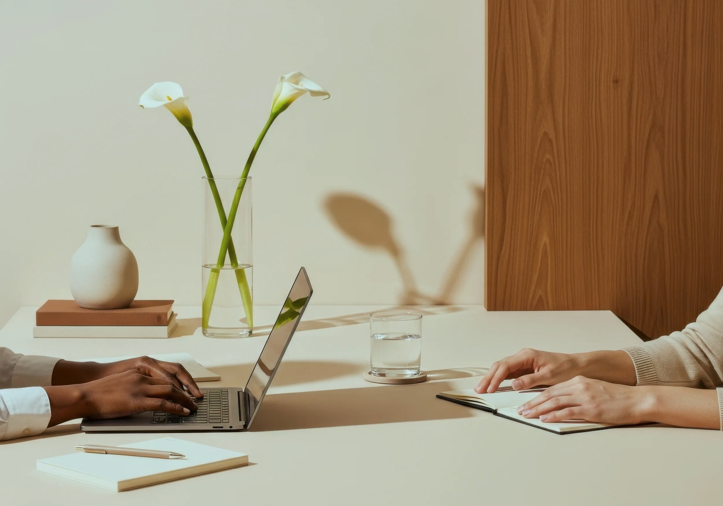 Two people working at a white table, one using a laptop and the other writing in a notebook, with a glass of water, a pen, and notebooks. On the wall behind are two white calla lilies in a glass vase and a small white ceramic vase, with a wooden panel on the right.