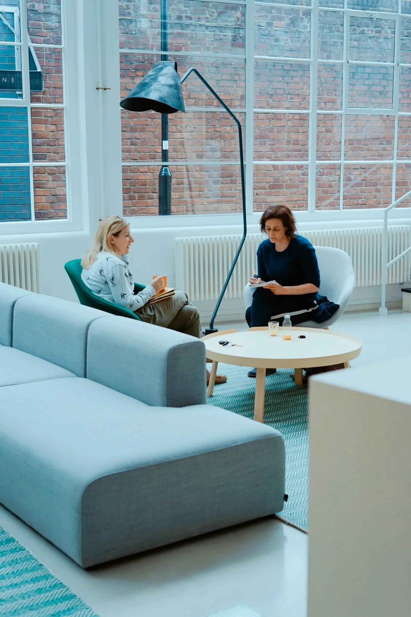 Two women sit in a bright, spacious room with large windows and brick walls. One woman, with blonde hair, is drinking from a mug, and the other woman, with dark hair, is taking notes. A floor lamp and a round coffee table with items are in the room.