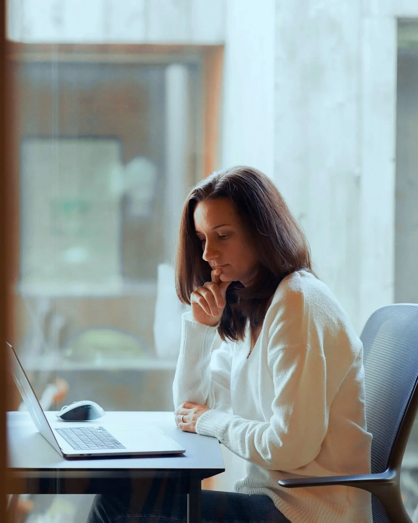 A woman with brown hair wearing a white sweater sitting at a desk in front of a laptop, looking thoughtful or focused.