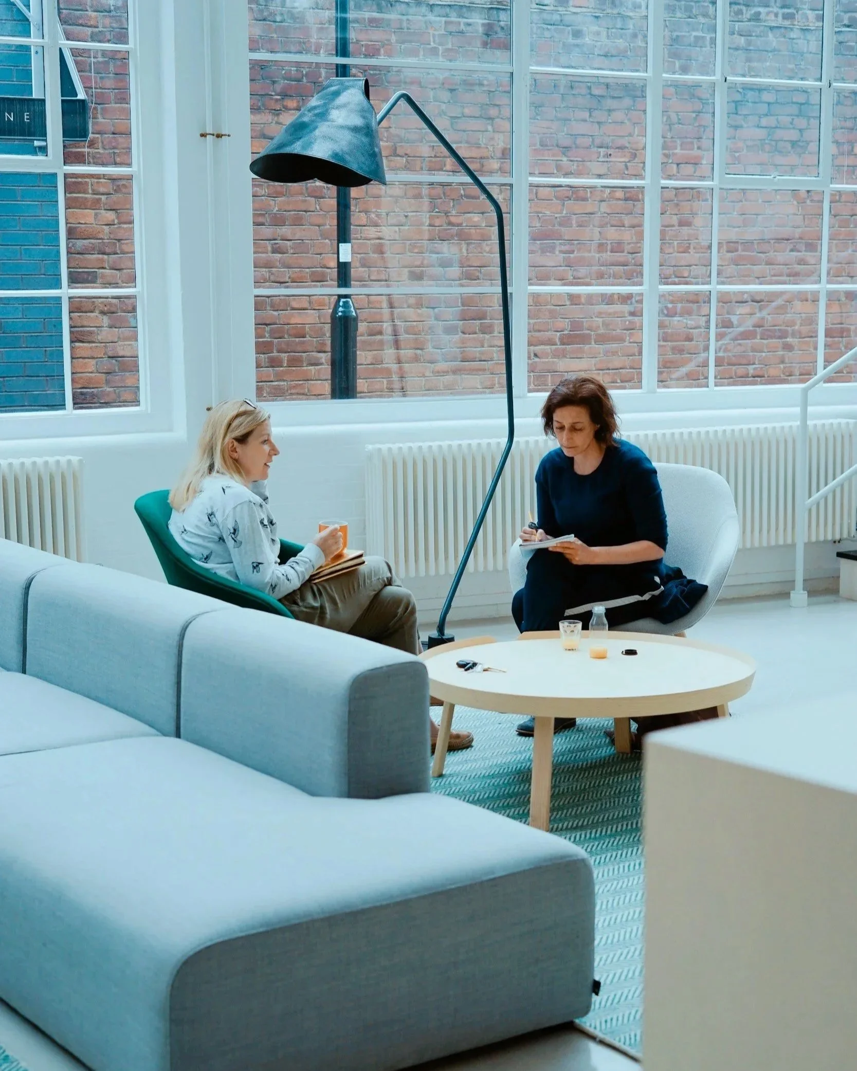 Two women sitting in a bright room with large windows and brick walls, one talking and writing in a notebook, the other listening, with a small round table with water bottles and a notebook in front of them.