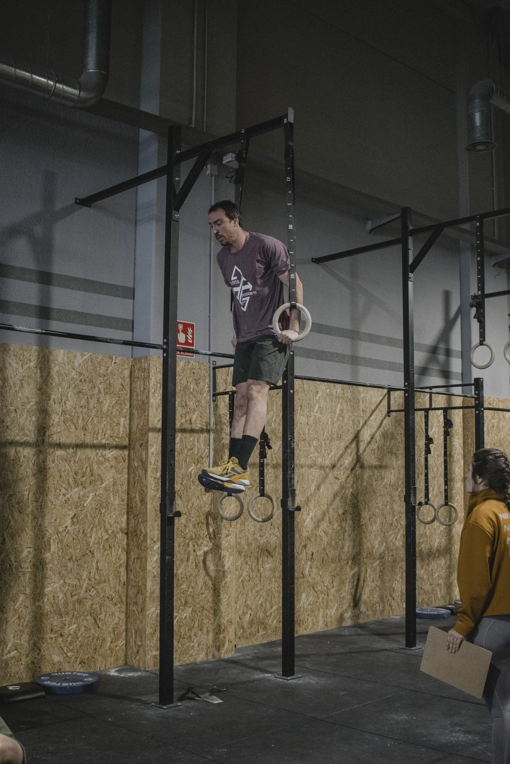 Man holding gymnastic rings while standing on a box in a gym with black padded floors and a wooden wall, with another person partially visible to the right.
