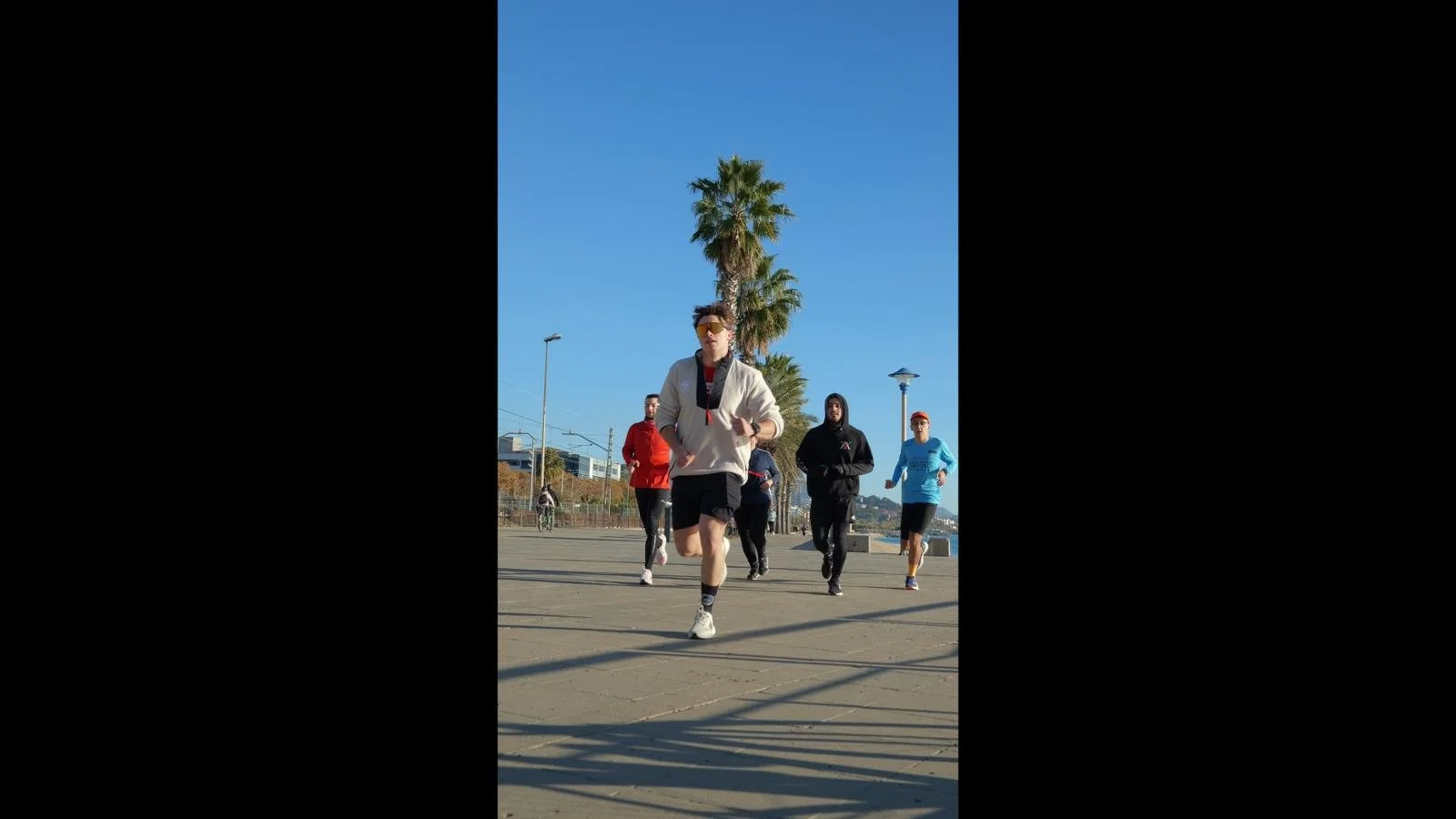 A group of five young men jogging on a sunny day in an outdoor park with palm trees, lampposts, and a clear blue sky in the background.