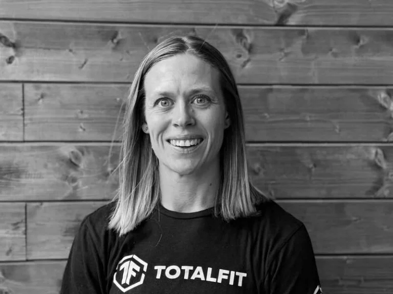 Black and white portrait of a woman with shoulder-length hair, smiling, wearing a T-shirt with 'TOTALFIT' logo, standing against a wooden background.