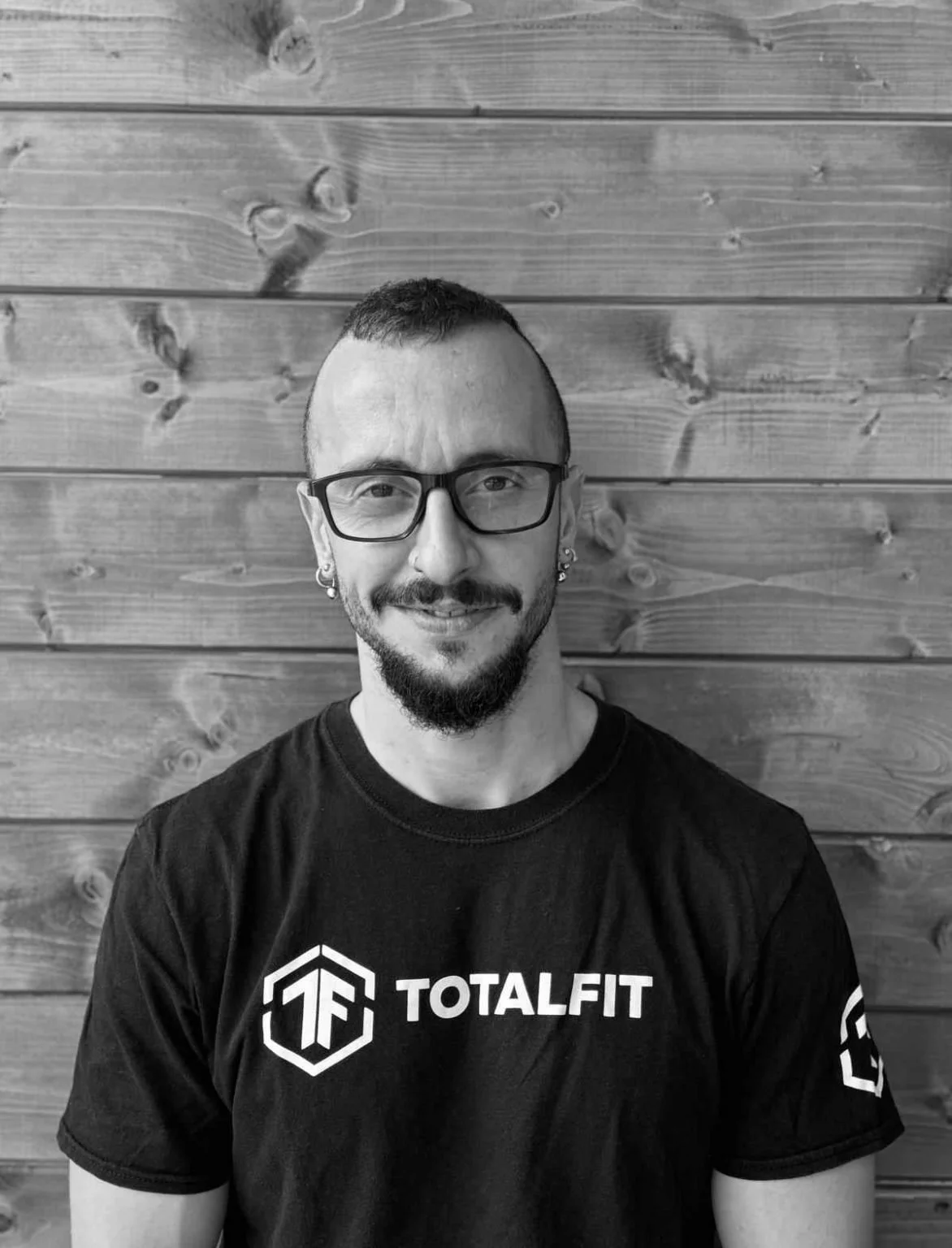 A man with glasses and earrings smiling, wearing a black T-shirt that says 'TOTALFIT' in front of a wooden wall.