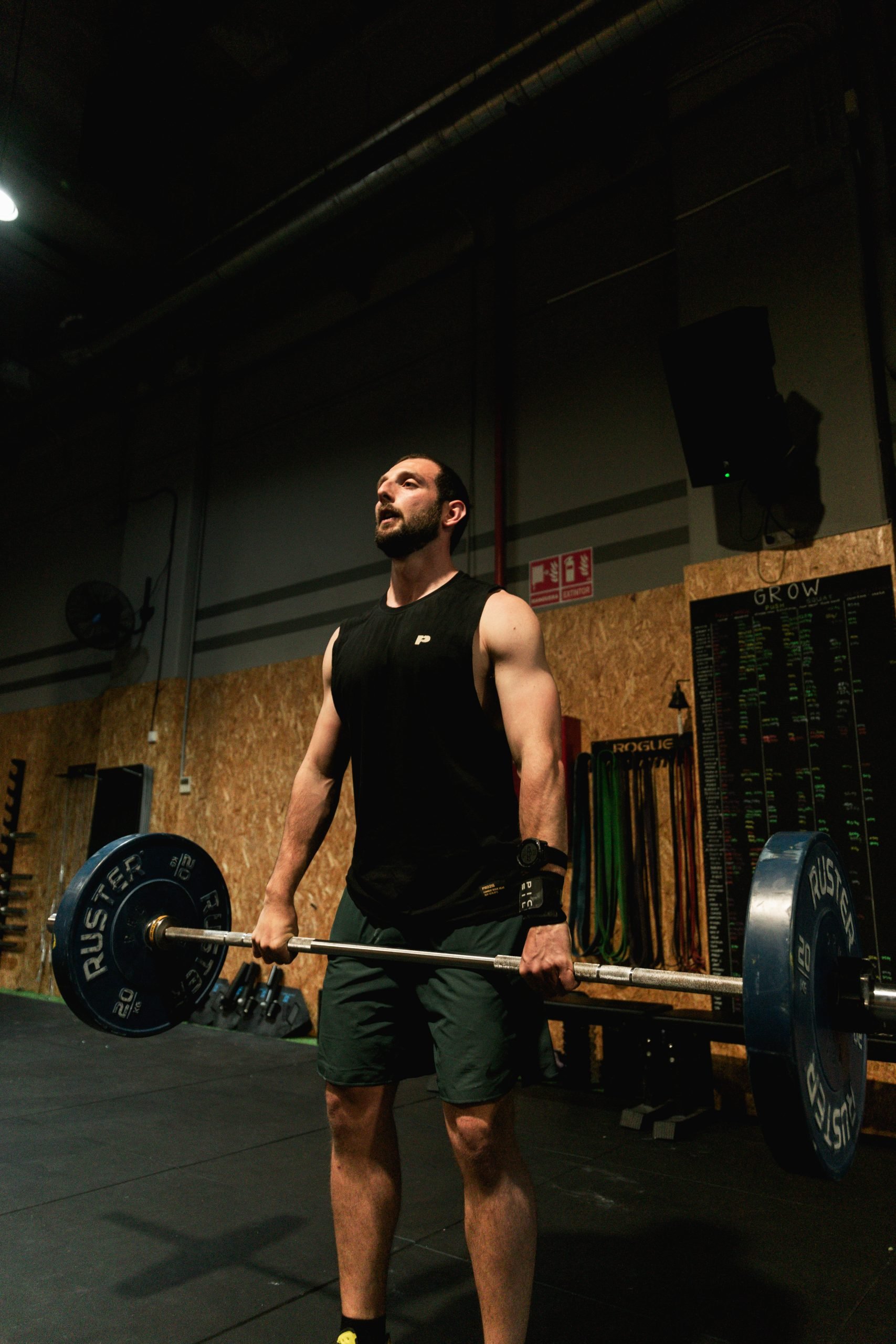 A man in a black sleeveless shirt and dark shorts lifting a barbell with weights in a gym. The background includes a wooden wall, a chart, and gym equipment.