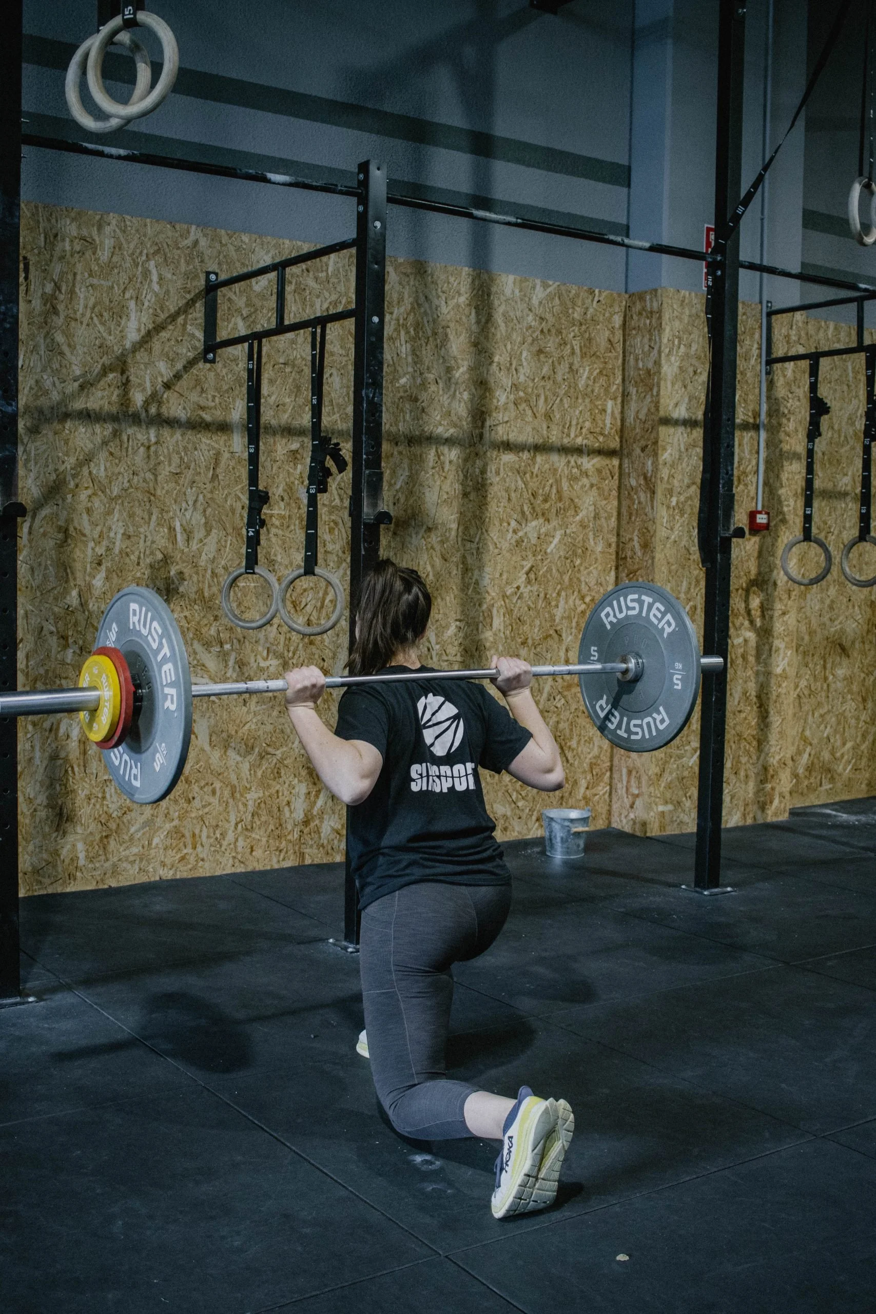 A person performing a squat exercise in a gym, holding a barbell with weights on their shoulders, kneeling on one knee on a black mat, with gym equipment and wooden wall in the background.