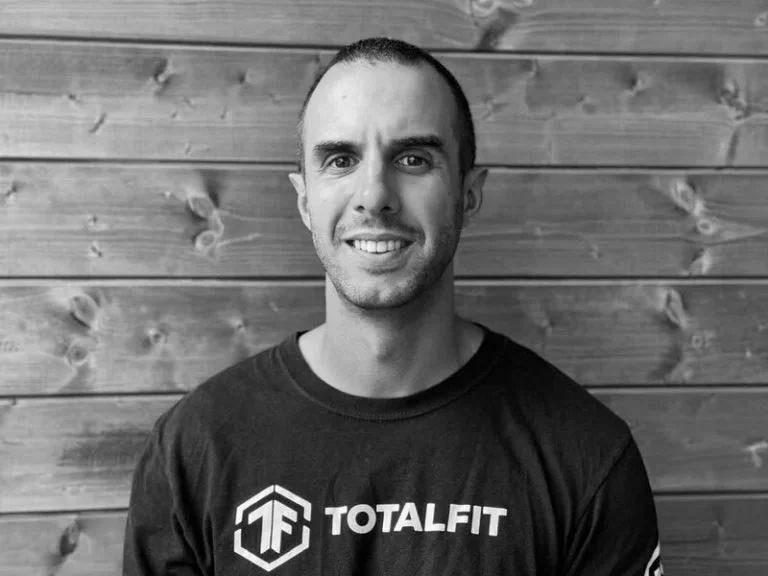 A smiling man with short hair and a beard wearing a black 'Total Fit' t-shirt, standing in front of a wooden wall.
