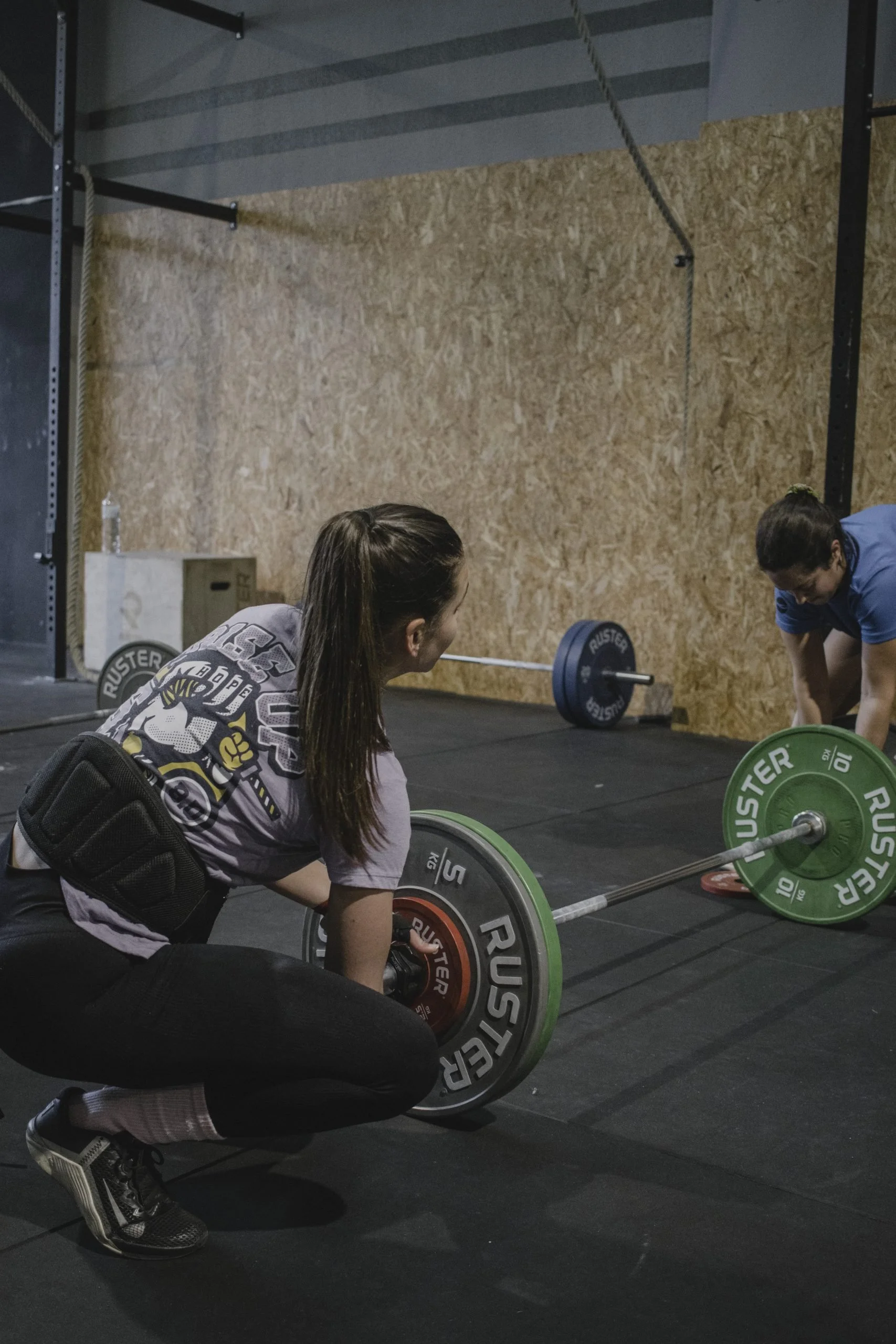 A woman with long dark hair in a ponytail, wearing a gray T-shirt with a graphic design, black pants, and sneakers, is in a gym setting performing a deadlift with a barbell loaded with green and black weight plates. Two other women are nearby, one on the floor and the other standing, assisting or spotting her. The gym has a black rubber floor, a portable wooden box, and a wooden wall with a horizontal metal bar and a climbing rope hanging from the ceiling.