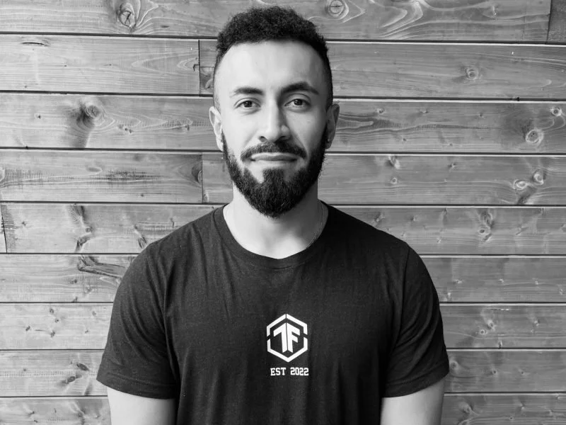 A man with a beard and short curly hair standing in front of a wooden wall, wearing a black T-shirt with a logo and text that reads "EST 2022."