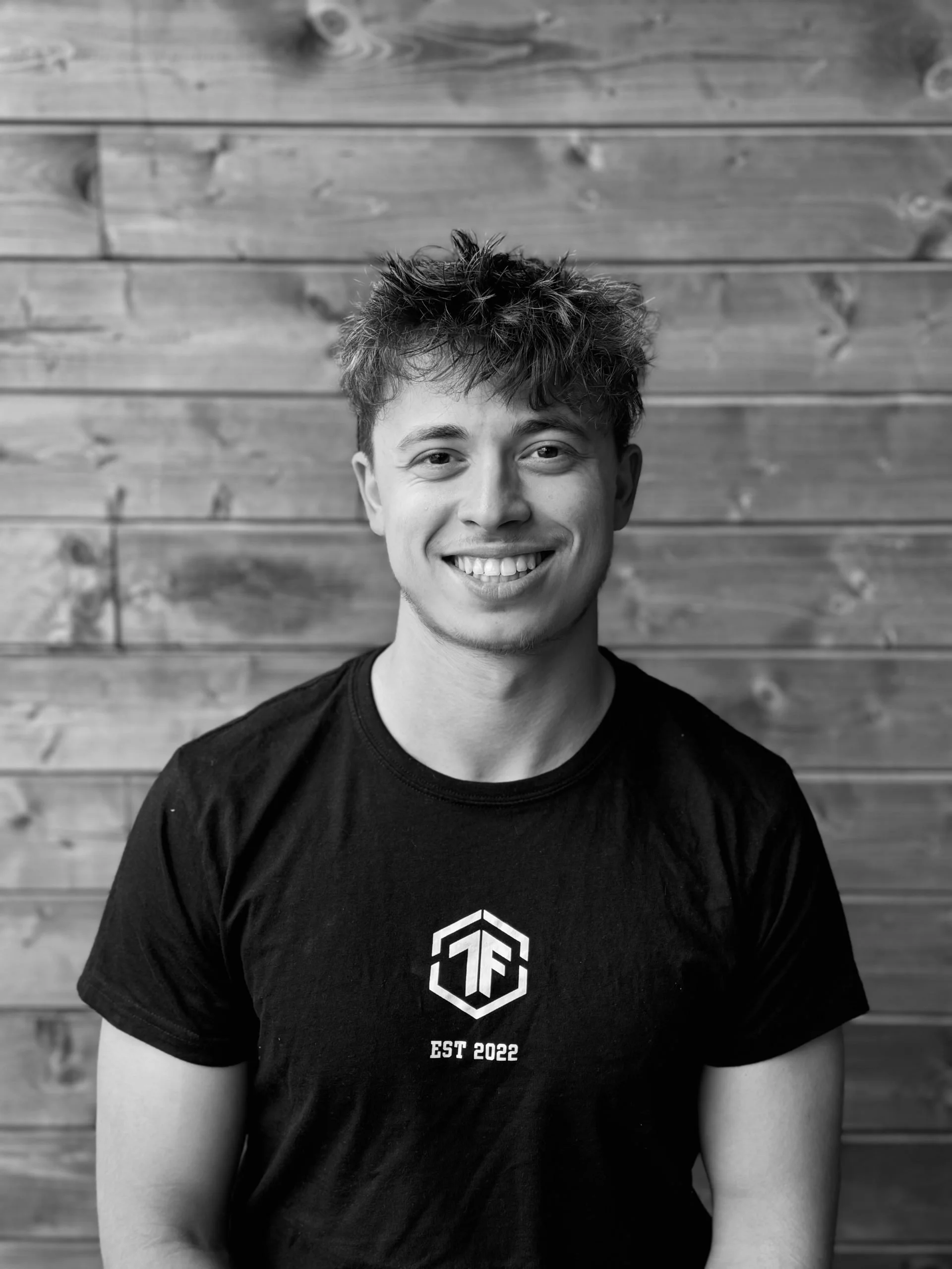 Black and white photo of a young man smiling, wearing a black T-shirt with a geometric logo on the chest, standing in front of a wooden wall.