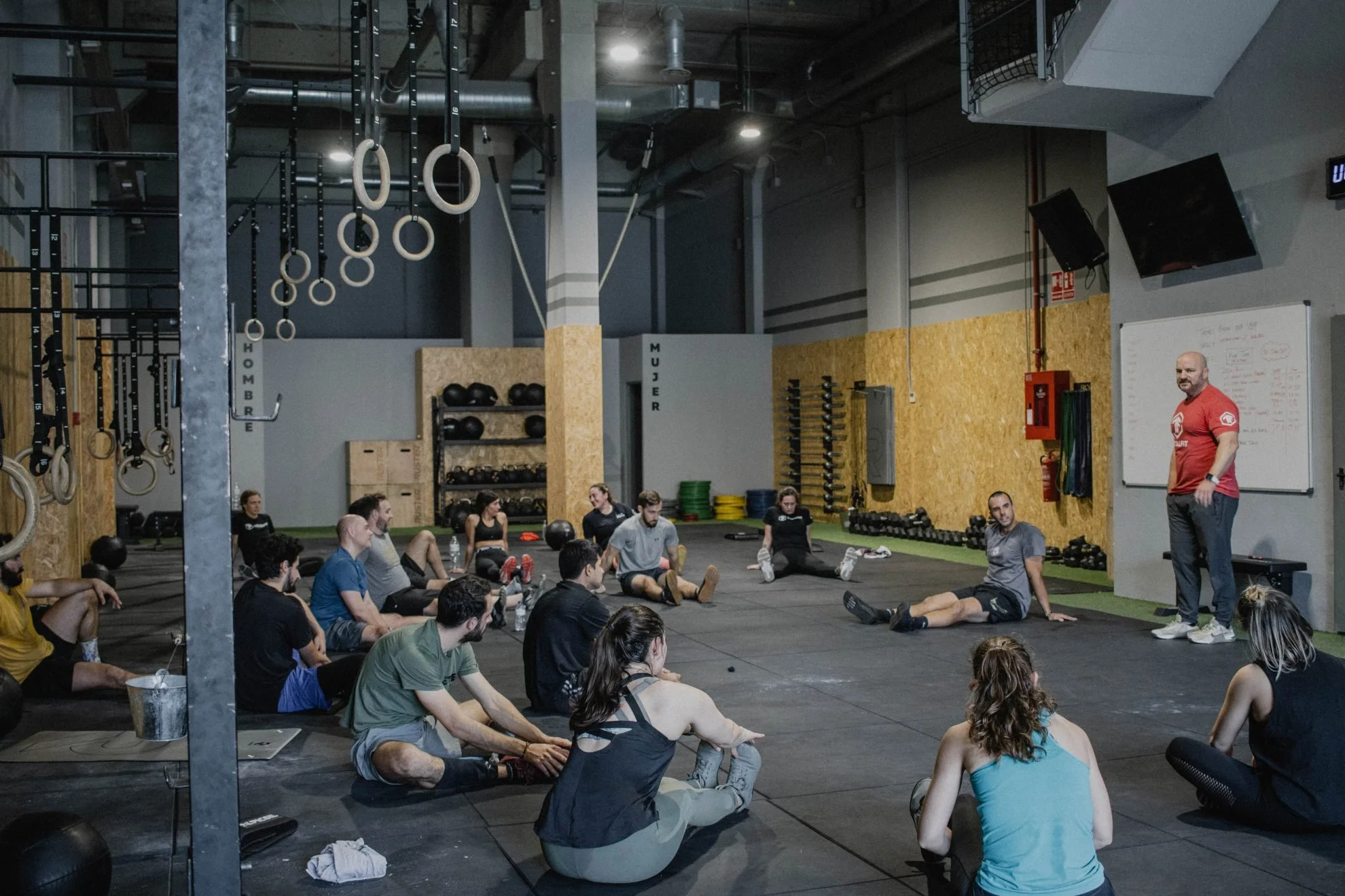 A group of people sitting on the floor in a gym, listening to an instructor who is standing near a whiteboard in front of them. Gym equipment like medicine balls, kettlebells, and weights are visible in the background.
