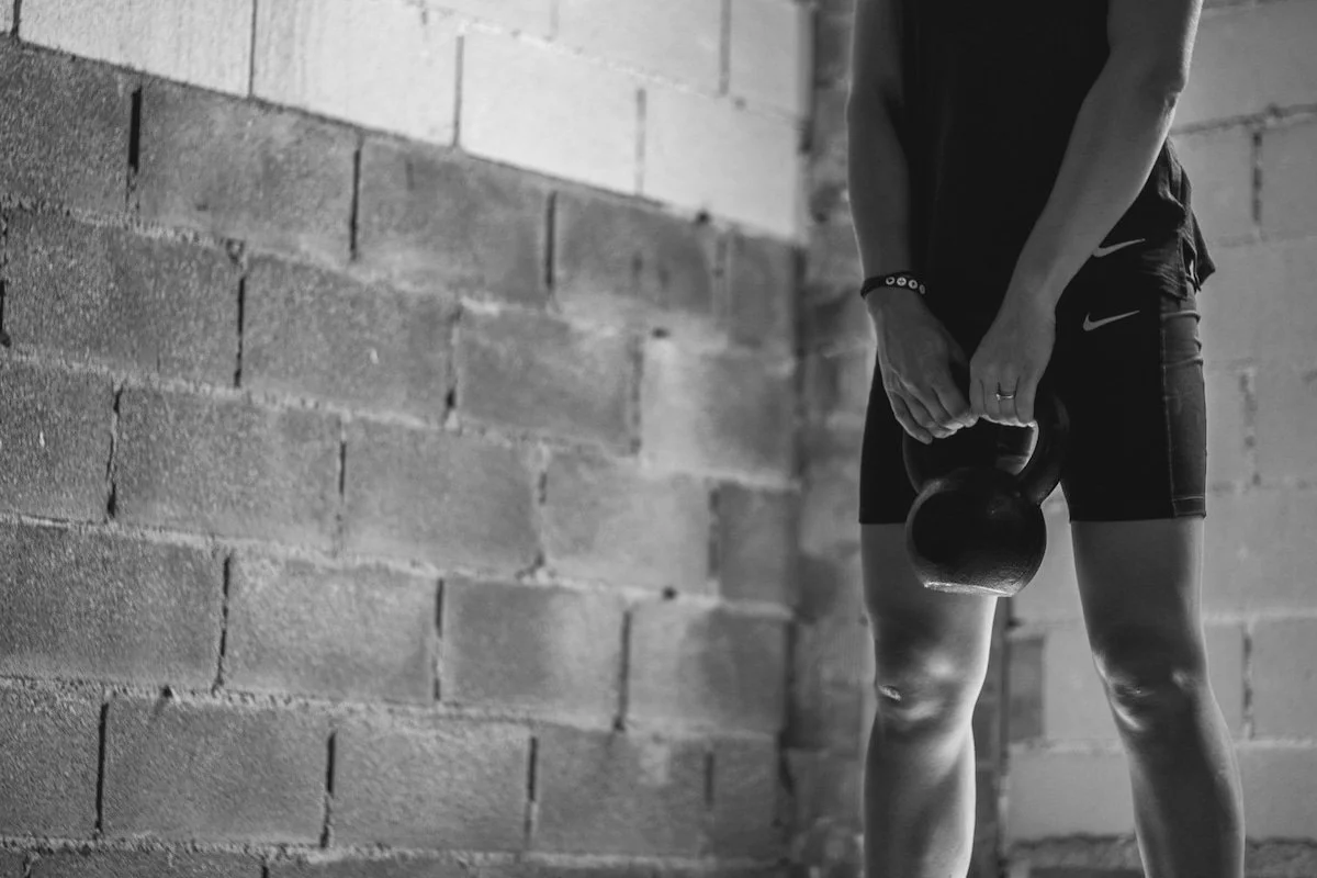 A person holding a kettlebell in a gym or workout space with a brick wall background.