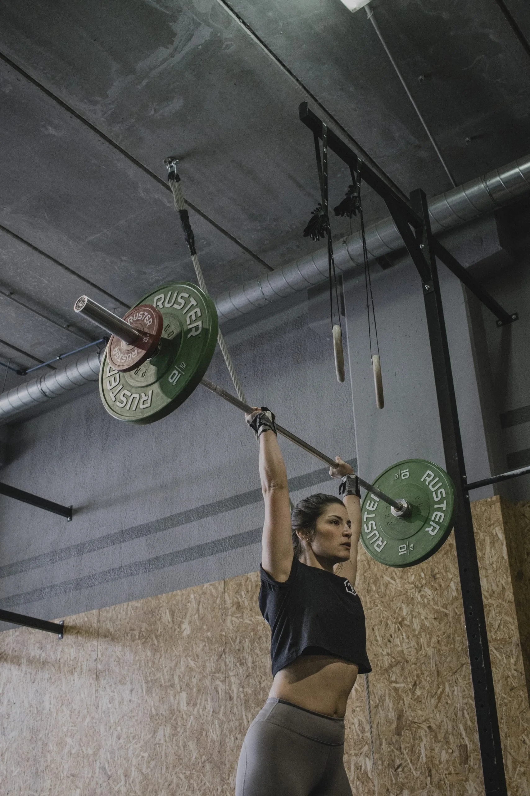 A woman is lifting a barbell with green weight plates overhead indoors, with her head tilted slightly back and wearing a black shirt and gray leggings.