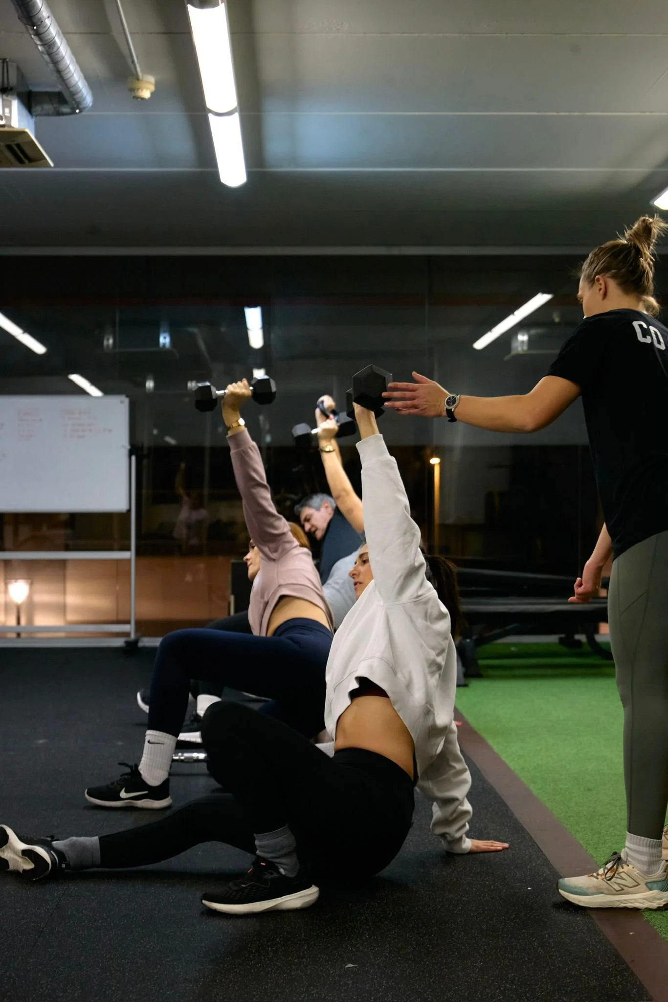 A group of people in a gym participating in a fitness class, with some lying on the floor and lifting dumbbells, and an instructor standing nearby. The gym has mirrors, bright lighting, and a mix of black and green flooring.