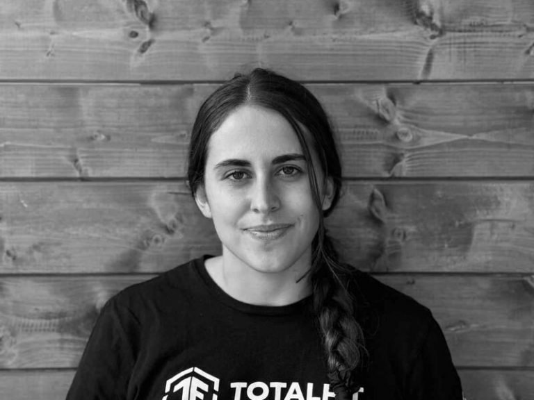 A young woman with long dark hair in a braid, wearing a black shirt with white text, smiling slightly, sitting in front of a wooden wall.