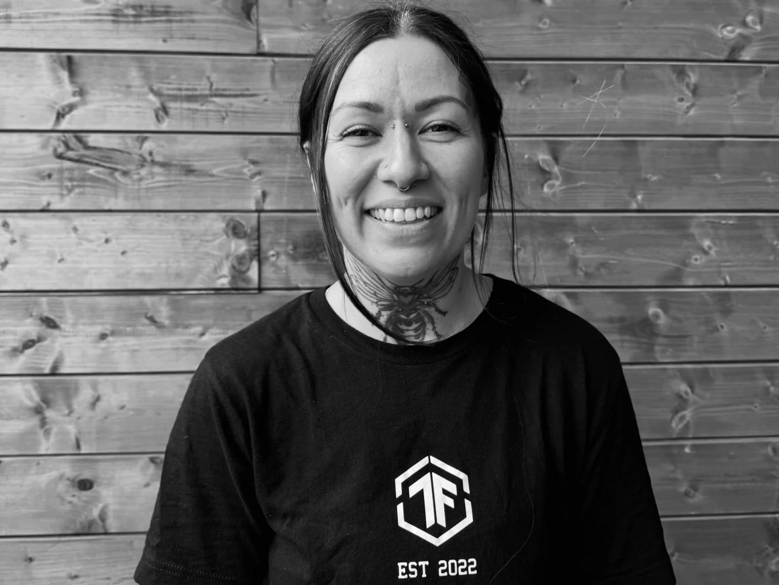 A woman with tattoos and piercings smiling in front of a wooden wall, wearing a black shirt with a white logo and 'EST 2022' printed below.