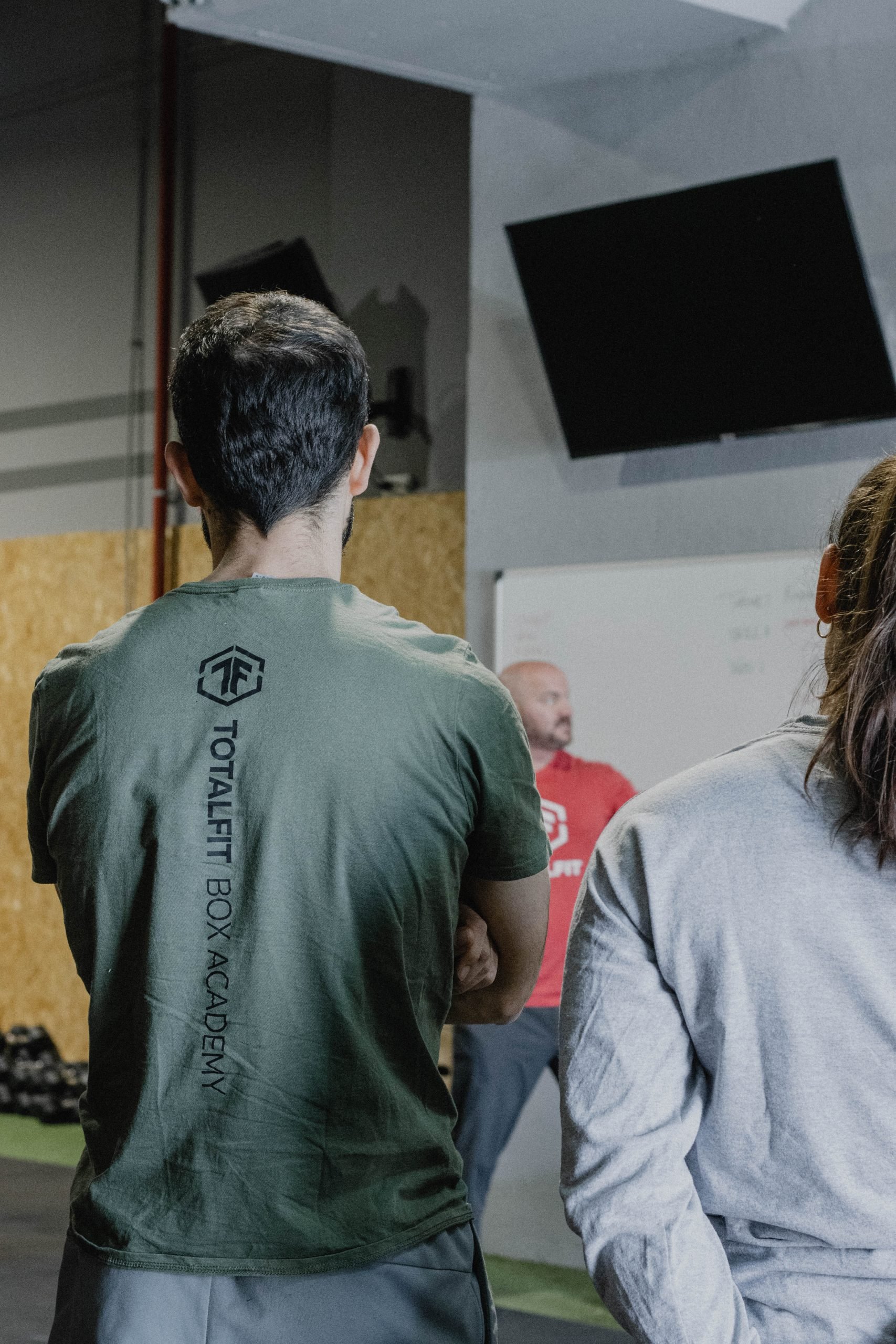 People attending a fitness class in a gym, facing forward towards a fitness instructor and a screen, with equipment on the side.