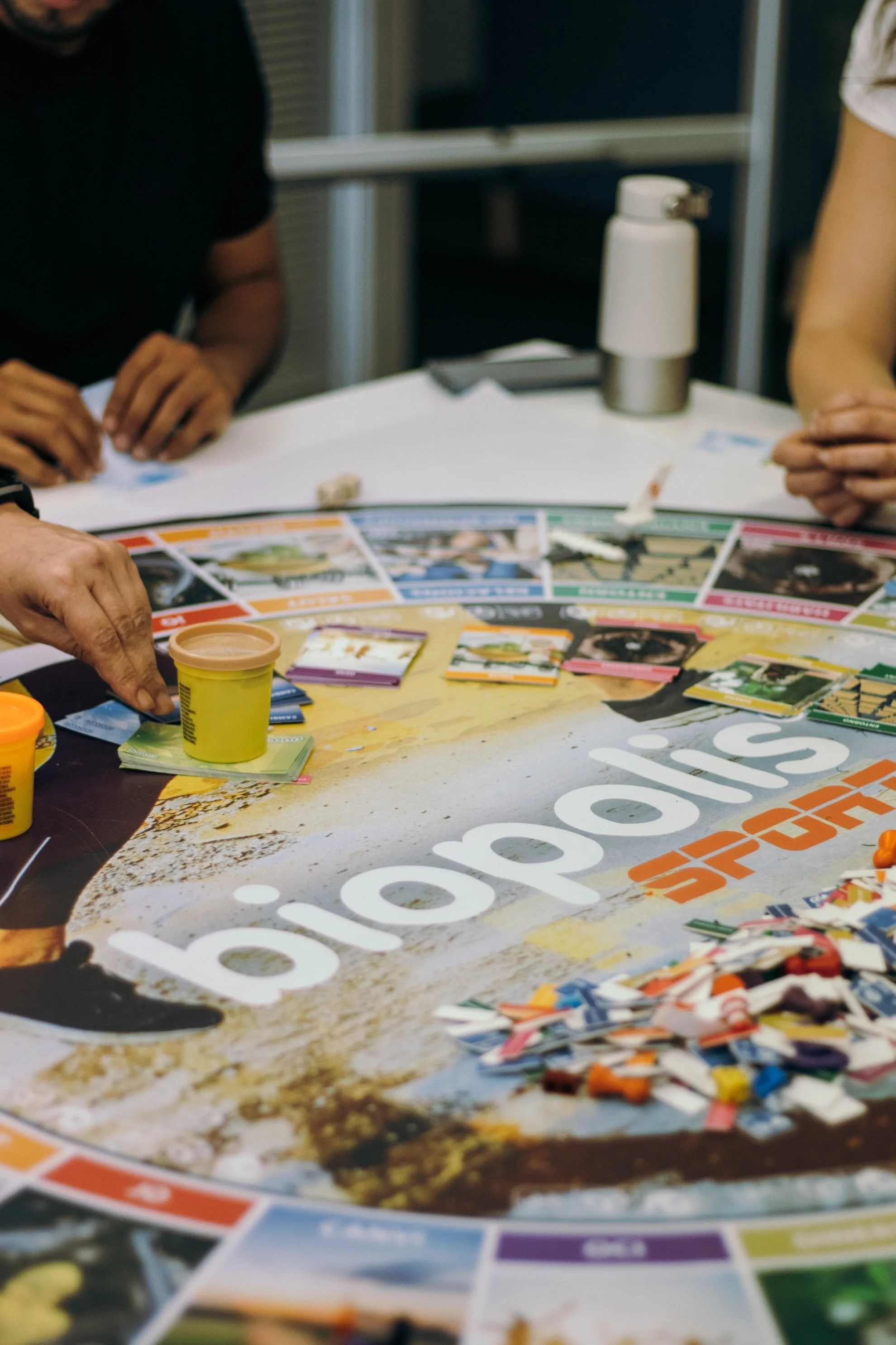 People playing a board game with colorful cards on a table, with a water bottle in the background.
